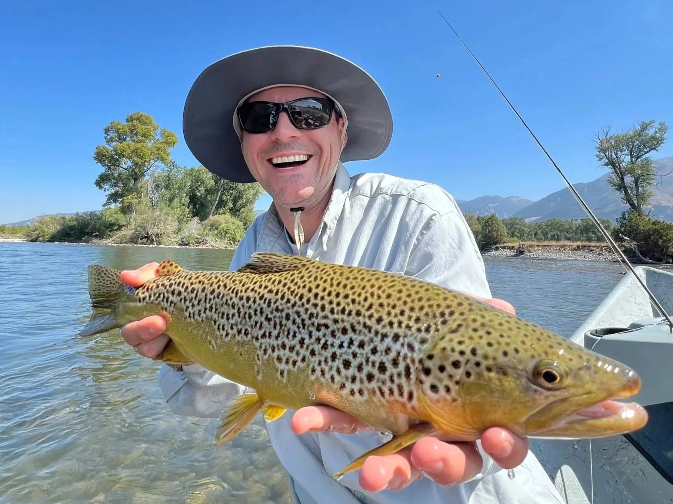 A smiling man wearing sunglasses and a wide-brimmed hat holding a freshly caught fish. The background shows a river, trees, and mountains under a clear blue sky.