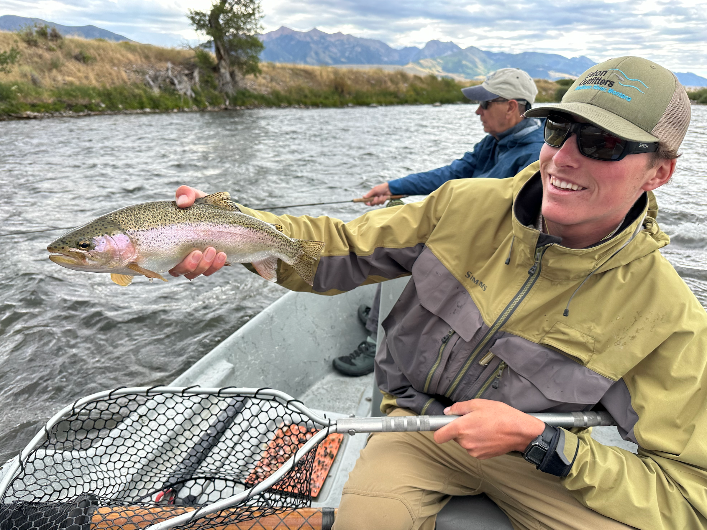 Two men fishing on a river in a mountainous area, one holding a caught fish, with fishing gear and a boat in the water.