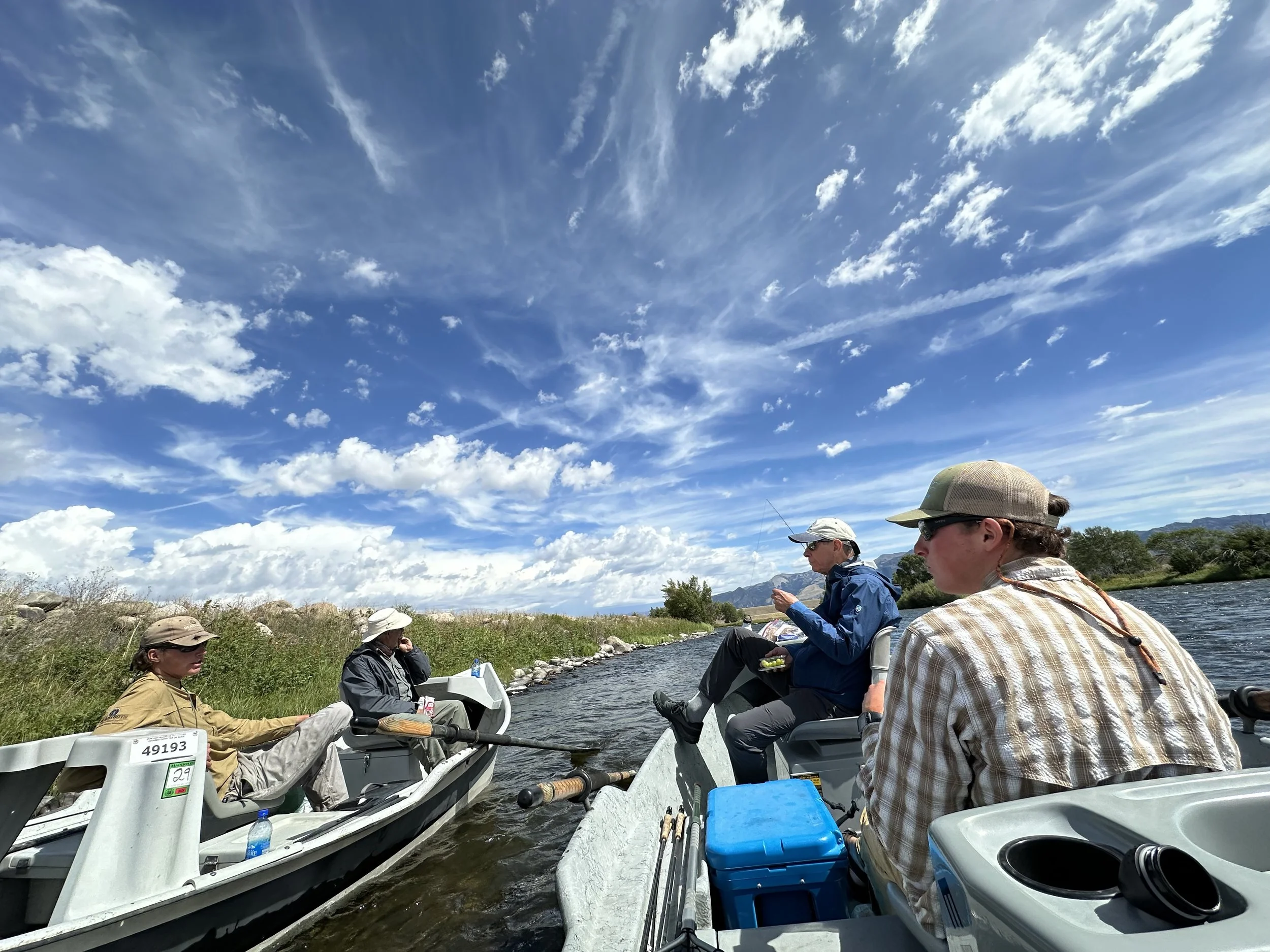 A group of people in boats on a river, fishing, under a partly cloudy sky, with mountains and trees in the background.