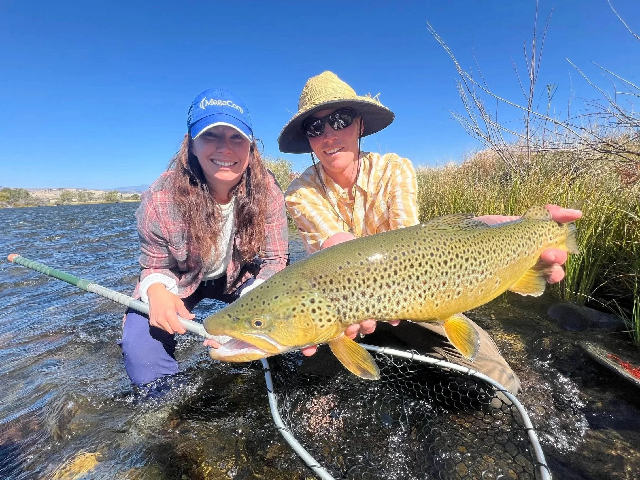 Two people in a river holding a large fish, blue sky, grass, and distant hills in the background.