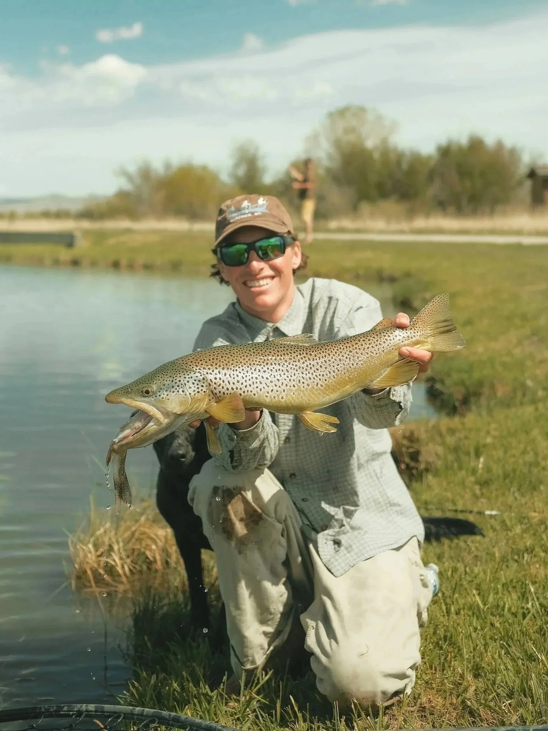 A man in sunglasses and a hat is kneeling at the edge of a pond, holding a large fish he caught.