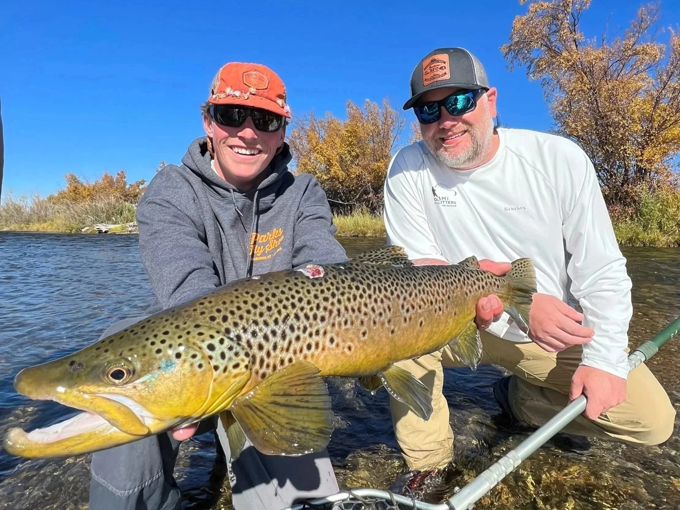Two men outdoors holding a large fish with colorful spots, near a body of water, under a clear blue sky with trees in fall foliage.