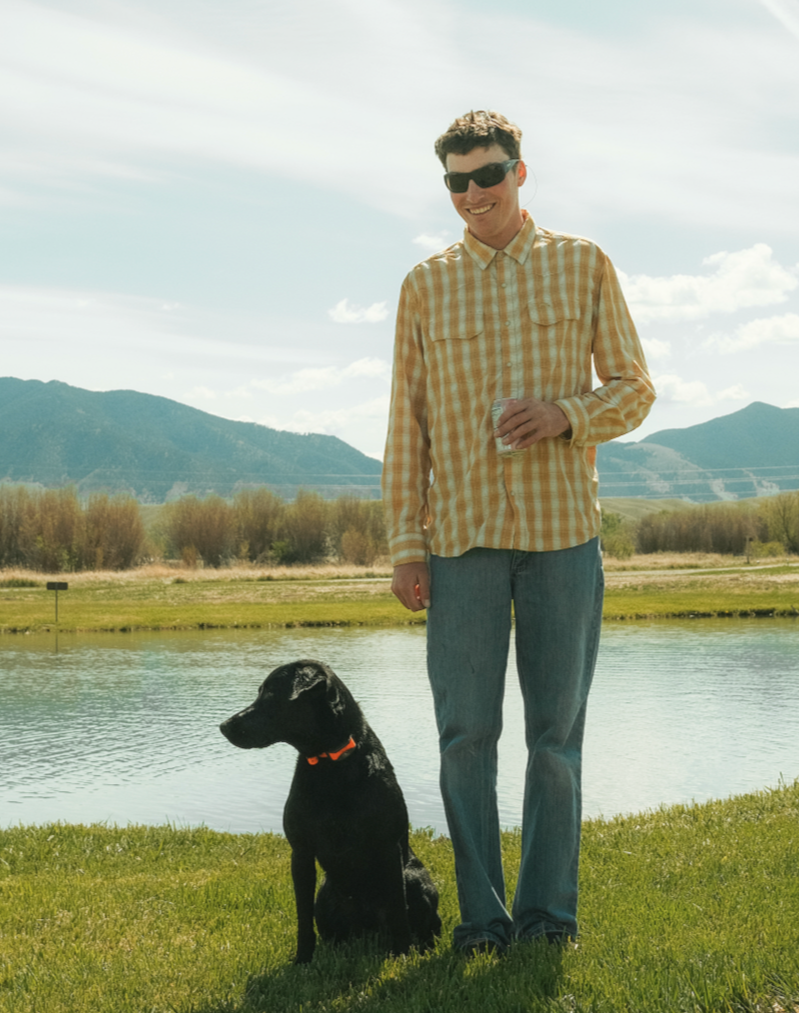 A young man wearing sunglasses and a yellow plaid shirt standing outdoors next to a black dog with an orange collar near a pond, with mountains and a partly cloudy sky in the background.