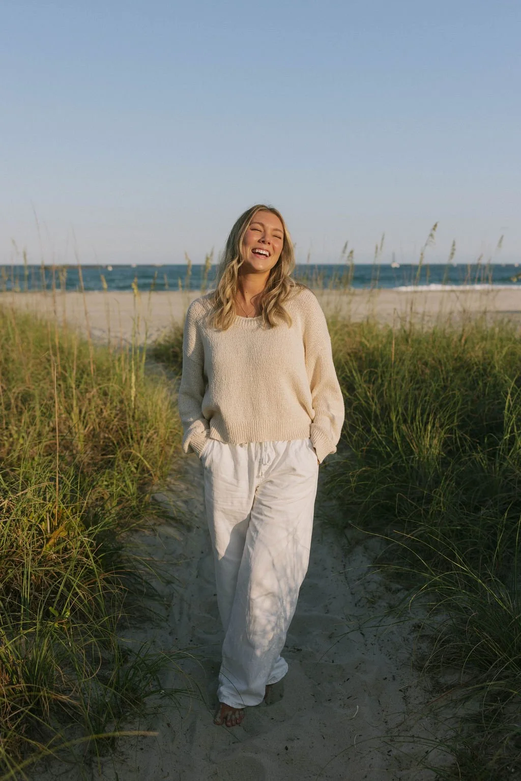 A young woman with blonde hair wearing a cream sweater and white pants standing on a sandy beach path, smiling with eyes closed, surrounded by tall grass, with the ocean and clear sky in the background.