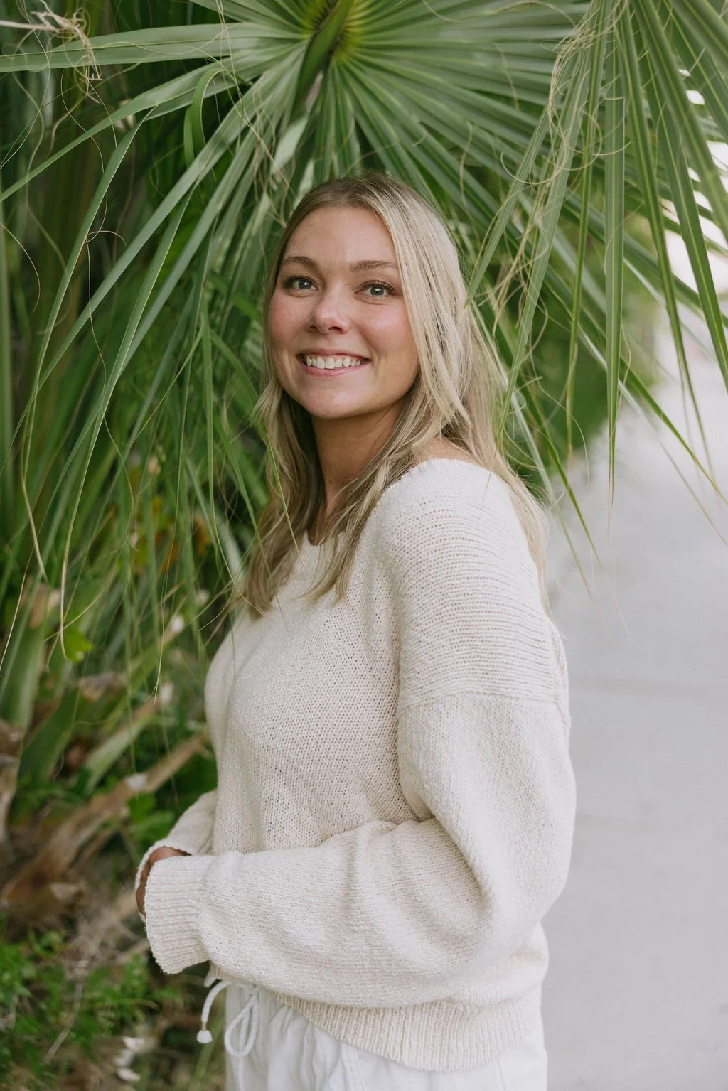 A woman with blonde hair smiling outdoors near large green tropical leaves.