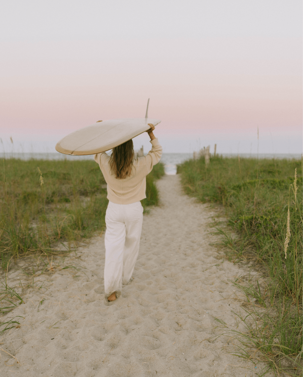 A woman walking on a sandy path towards the beach carrying a surfboard, surrounded by green grass and under a pastel-colored sky.
