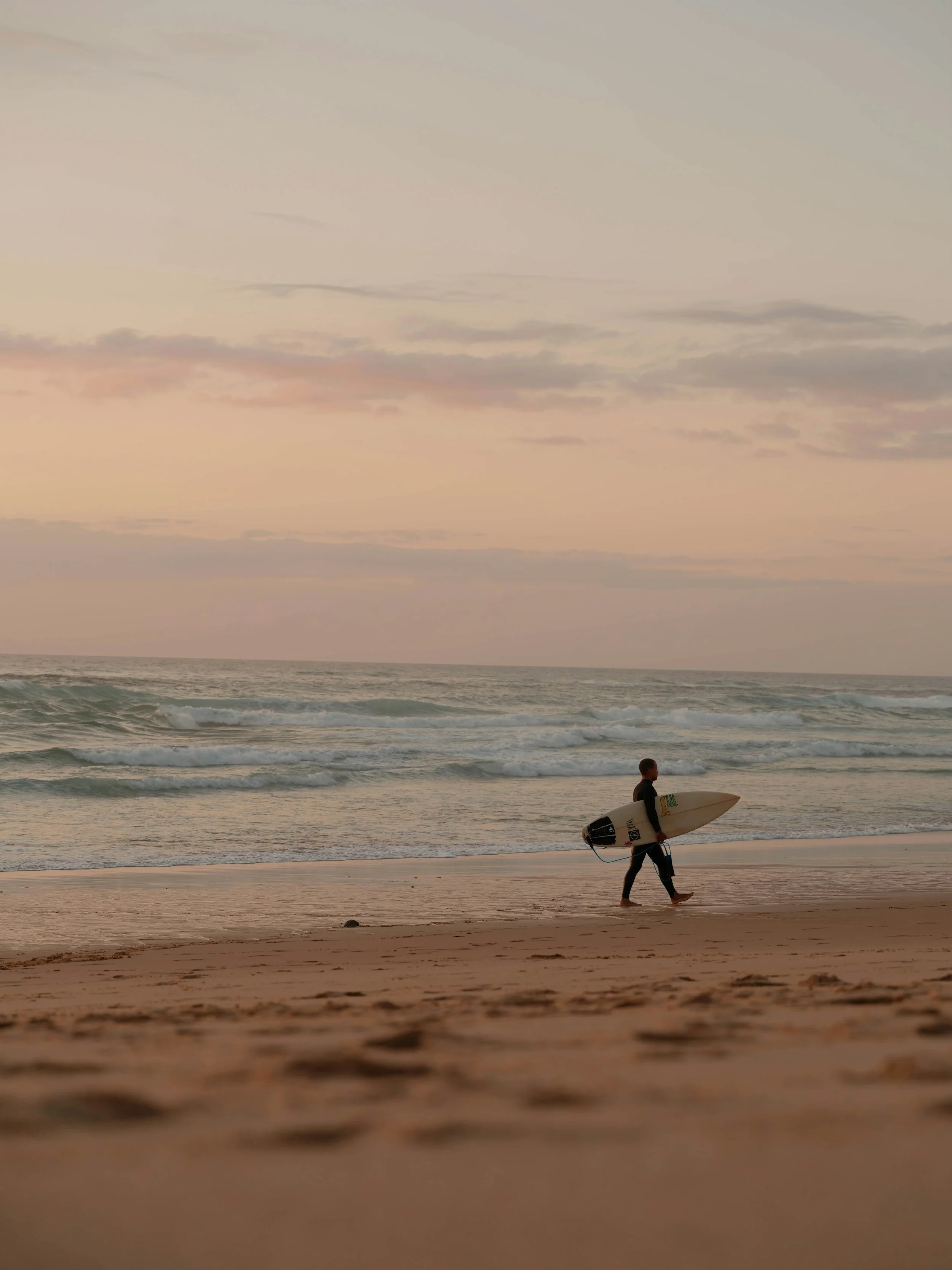 A person walking on a beach holding a surfboard, with ocean waves and a cloudy sky at dusk or dawn.