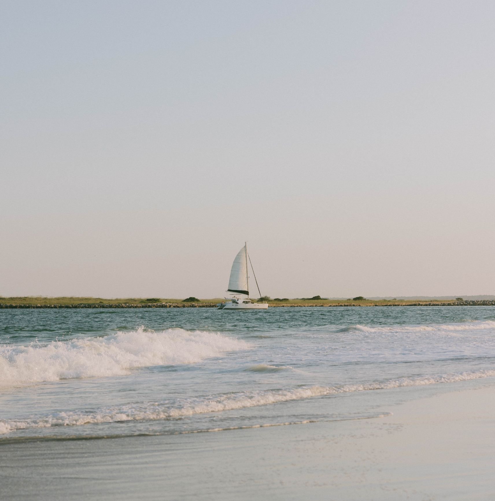 Sailboat on the water near a sandy beach with grassy land and clear sky in the background at sunset.