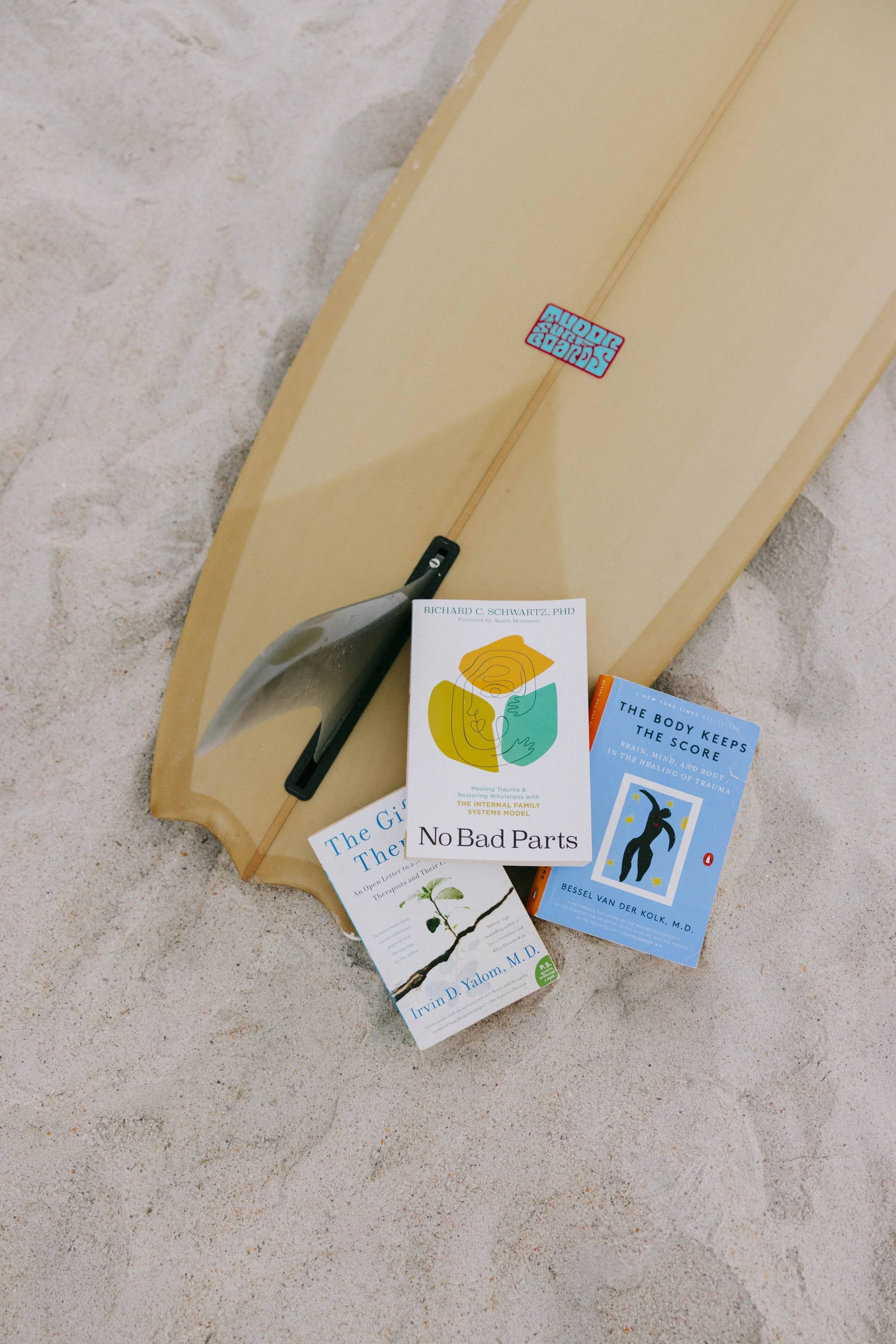 Various books and magazines placed on a surfboard on a sandy surface, with a golf club leaning against the surfboard, in a casual arrangement.