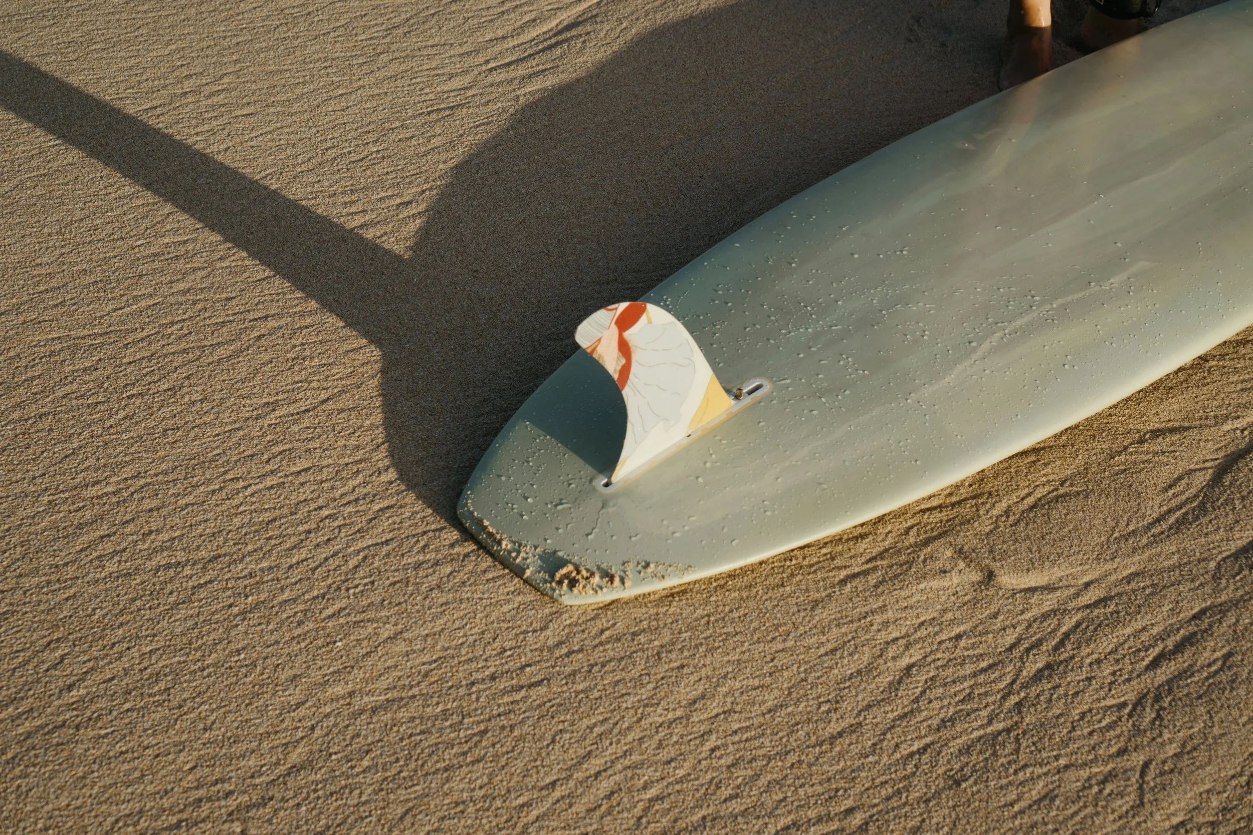 A surfboard lying on wet sand with water droplets on its surface, sun casting a shadow on the sand.
