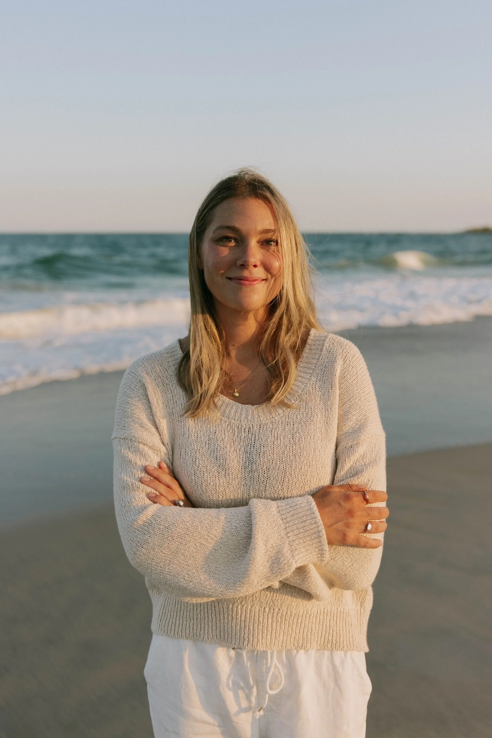 A woman with light brown hair and a gentle smile stands on a beach with waves in the background, wearing a cozy beige sweater and white pants, with arms crossed, during sunset.