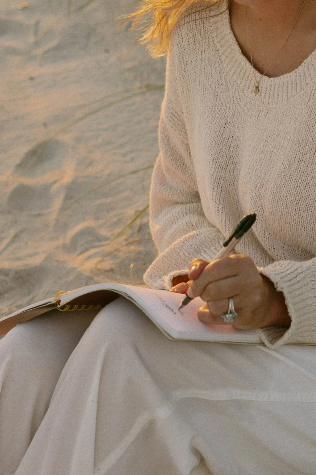 Person wearing a beige sweater and white skirt, writing in a notebook at the beach during sunset.