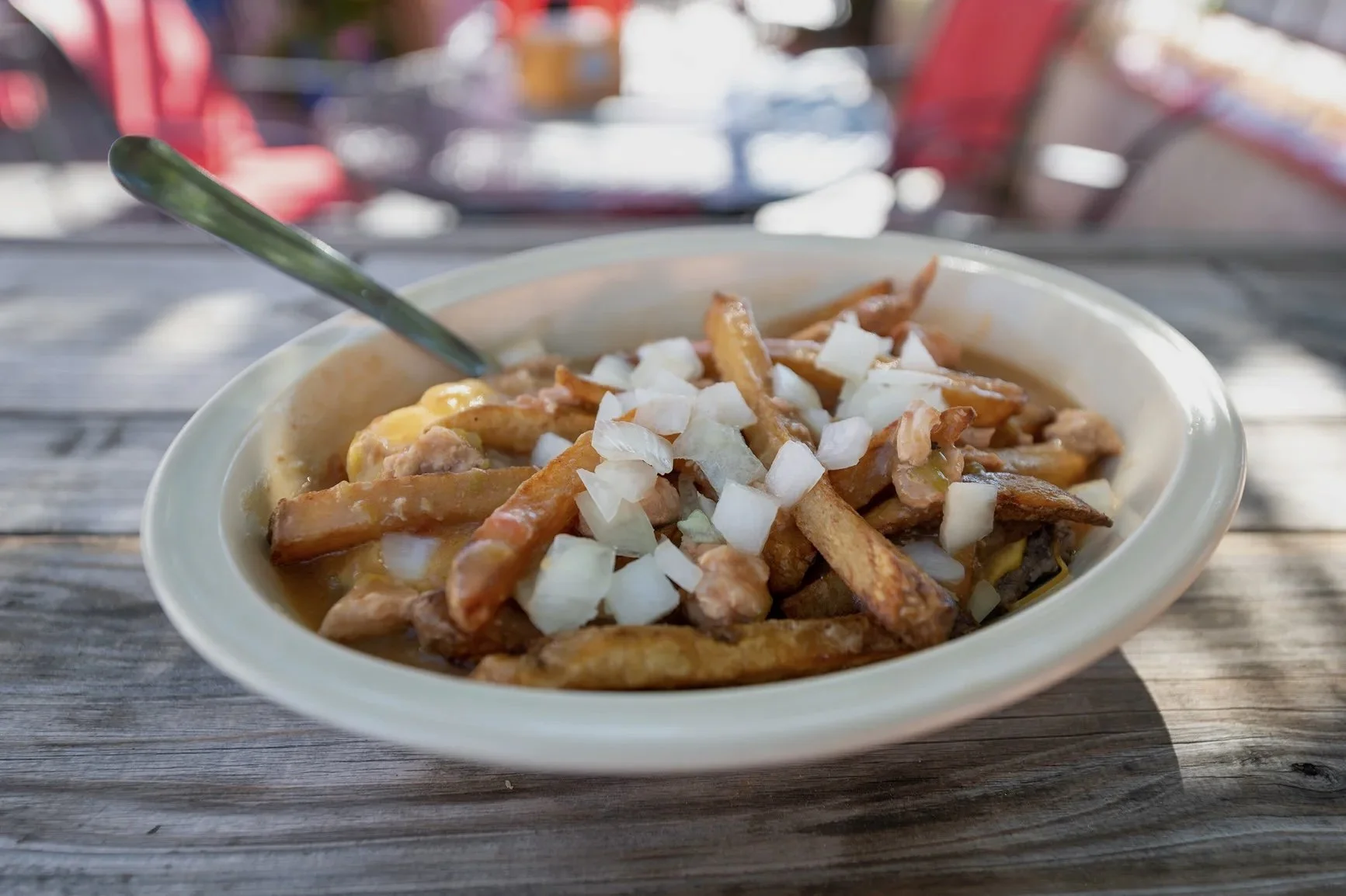 A bowl of poutine topped with chopped onions on a wooden table at an outdoor dining area.