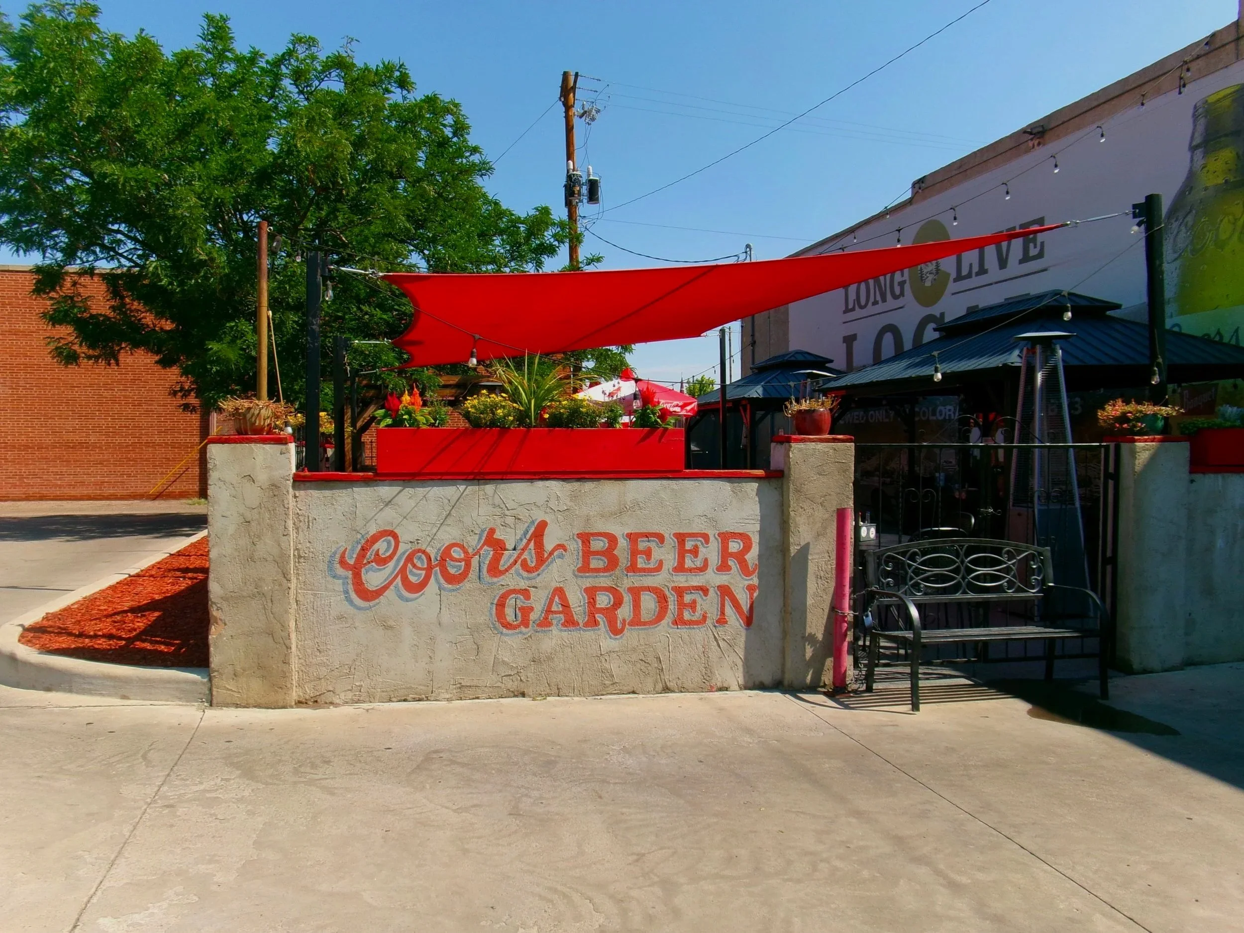 Outdoor restaurant or bar with a white stucco wall, painted with red lettering that reads 'COSTA BIER GARDEN' and a red fabric canopy shading the seating area. There are potted plants, black patio heaters, and string lights in the scene. A large gree