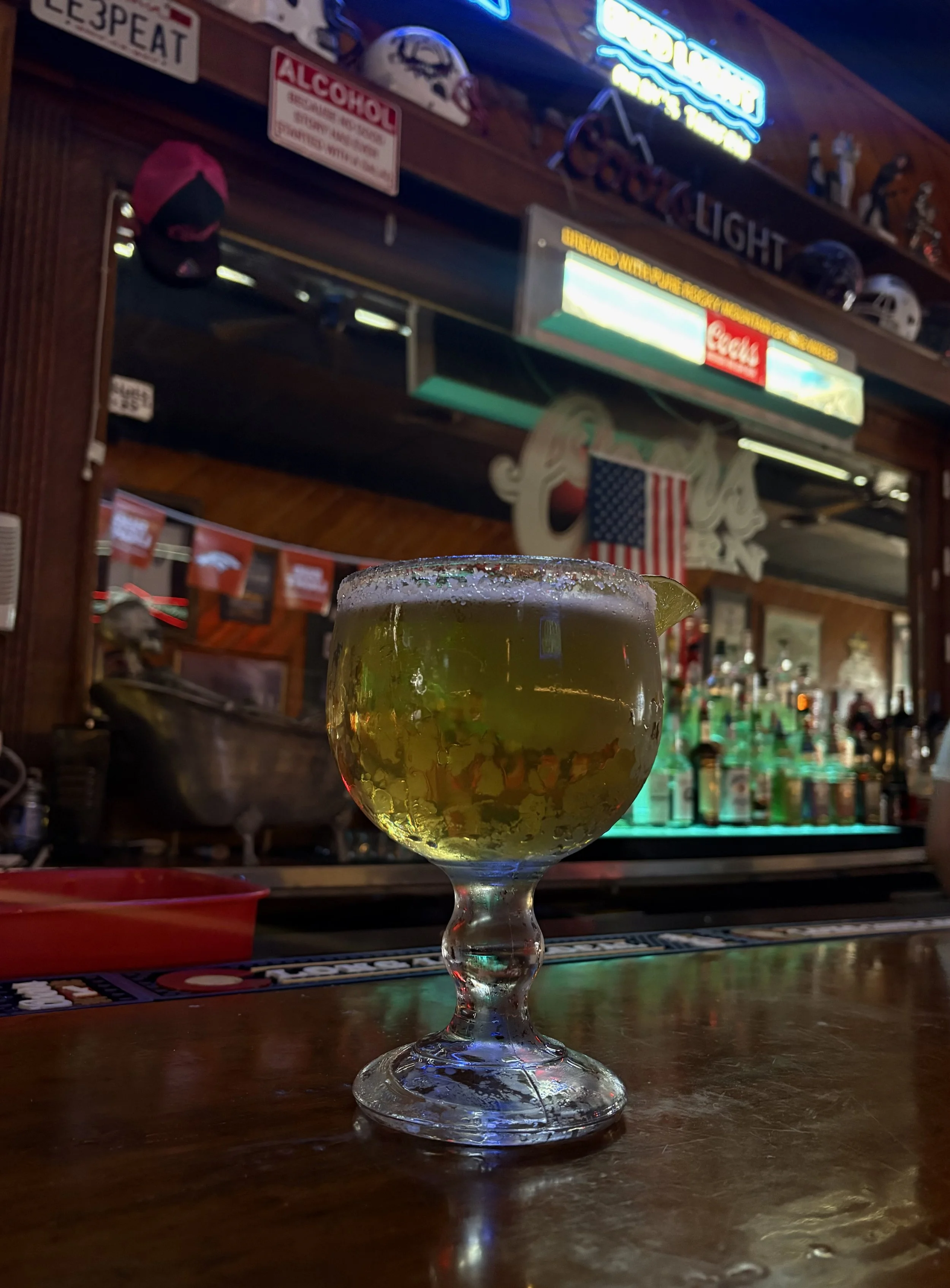 A margarita cocktail with a salted rim and lime wedge, sitting on a bar counter in front of a neon-lit bar with sports helmets, memorabilia, and an American flag in the background.