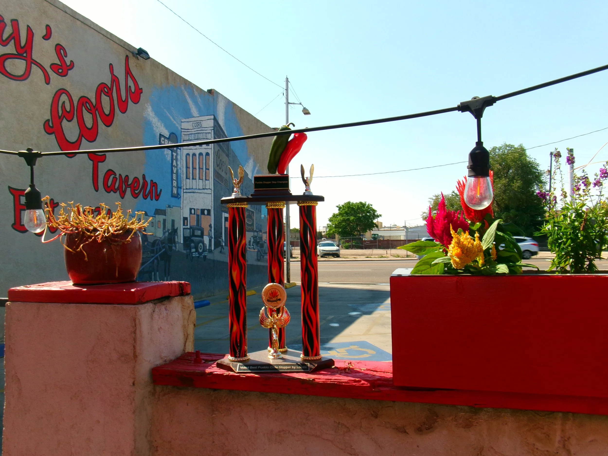 A trophy with red and black flames design and gold accents, placed on a red platform outdoors, with colorful flowers nearby, against a backdrop of a mural and a clear sky.