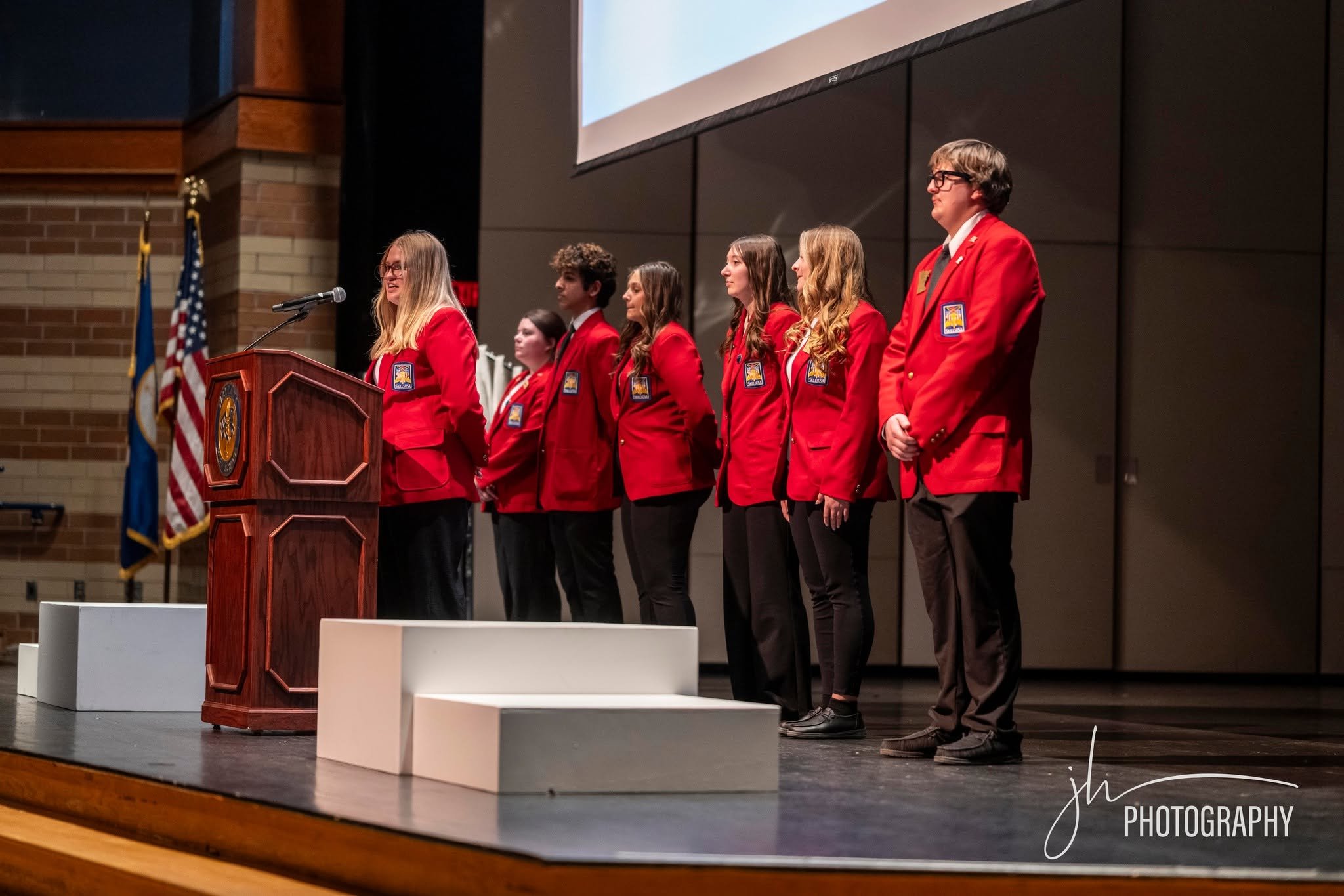 A group of seven young people in red blazers standing on a stage, with a podium and flags in the background, appearing to participate in a formal event or ceremony.