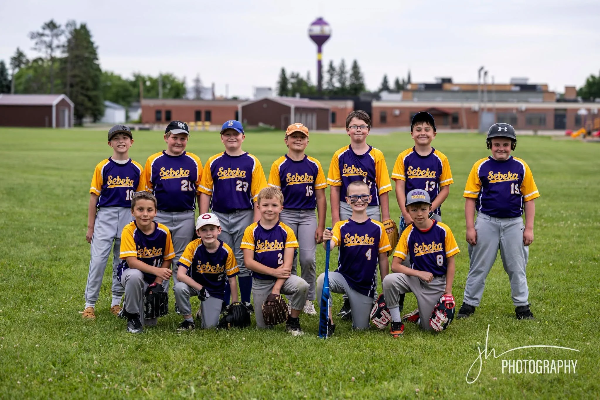 A group of young boys in baseball uniforms pose for a team photo on a grassy field. They are smiling and standing together, some holding baseball gloves and bats, with a few kneeling in front. The background features an open field with buildings and 