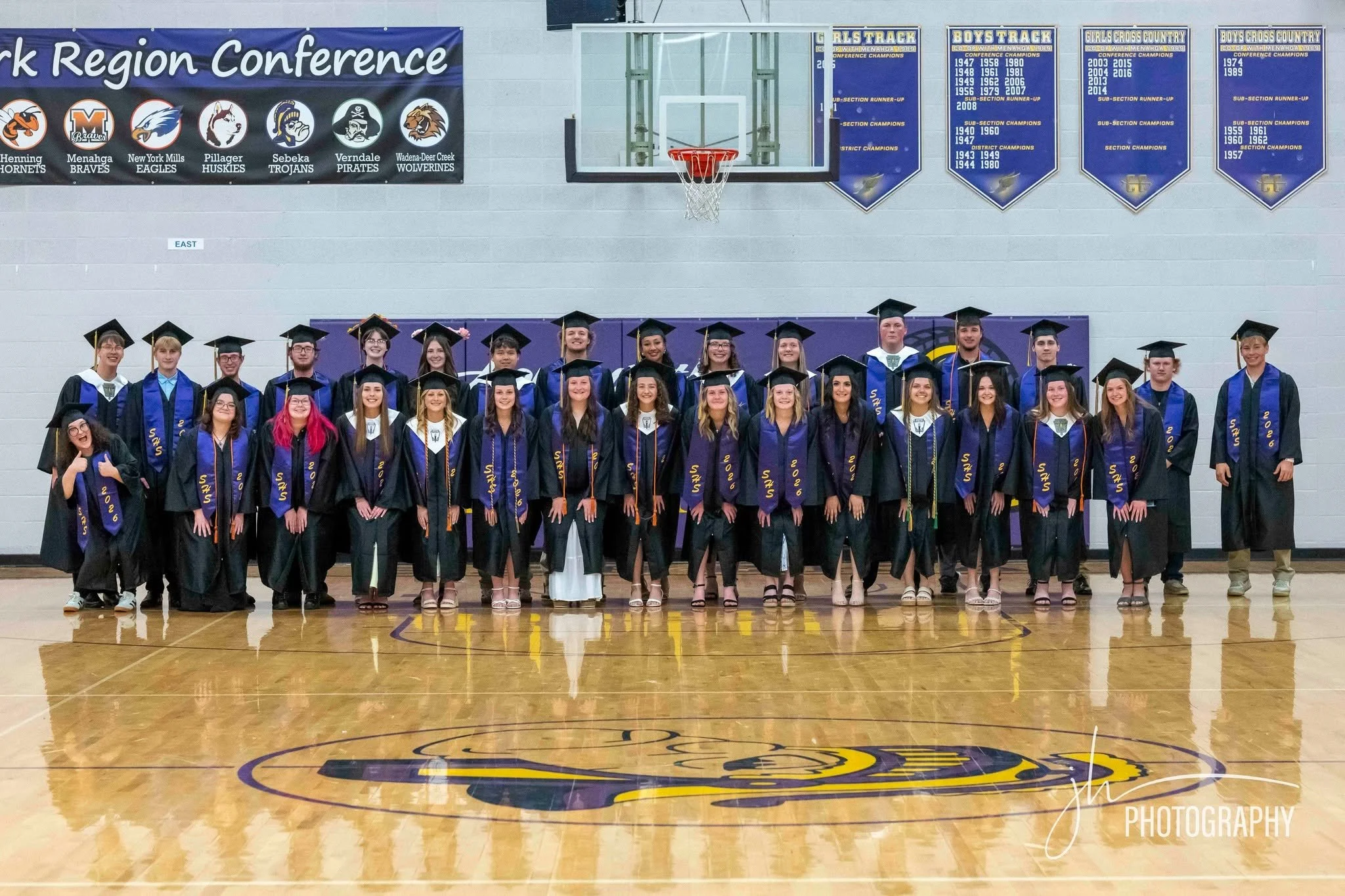 Group of high school graduates dressed in caps and gowns posing for a photo inside a gymnasium with banners and a basketball hoop in the background.