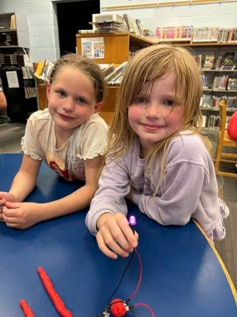 Two young girls sitting at a table in a library, smiling at the camera, with a science experiment setup in front of them.