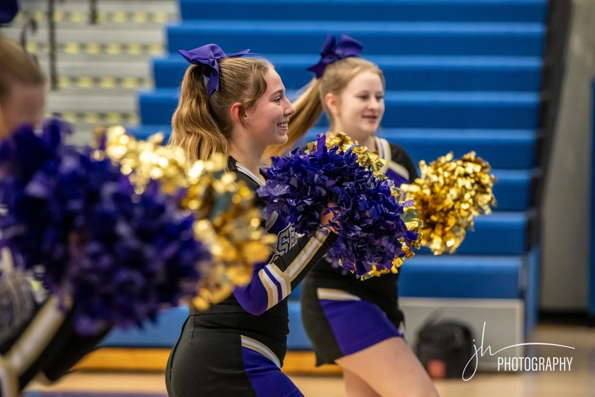 Cheerleaders in purple and black uniforms holding purple and gold pom-poms, standing in a gymnasium with blue bleachers in the background.