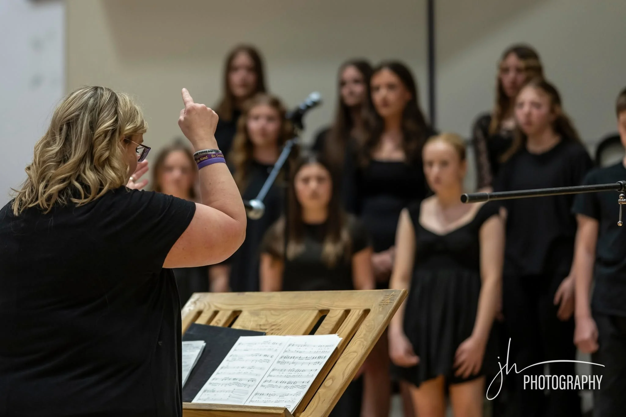 A woman with blonde hair and glasses conducting a choir rehearsal, with sheet music on a wooden music stand, and multiple women standing in the background.