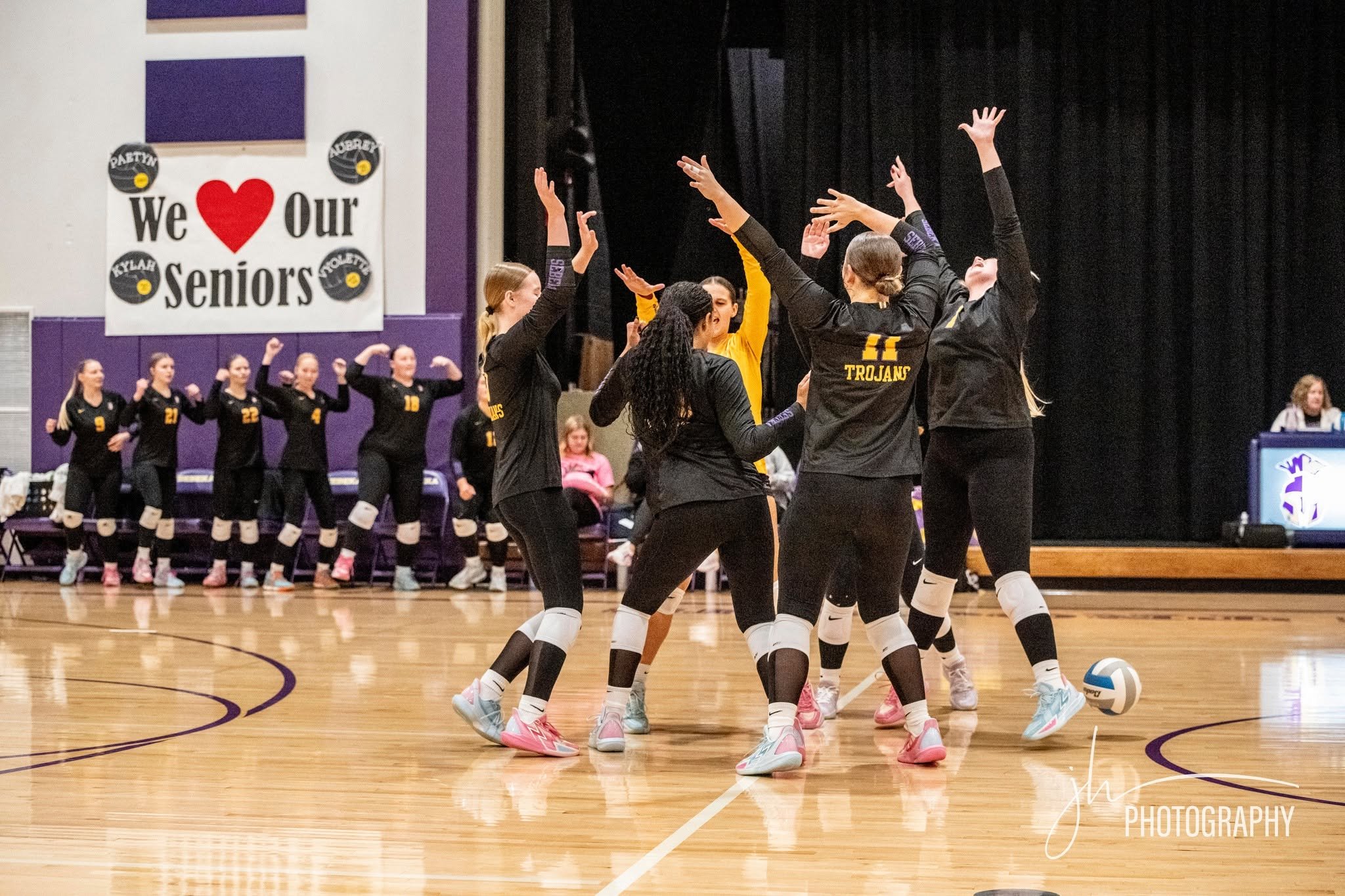 Volleyball team practicing on the court with a large sign that says "We ❤️ Our Seniors" on the wall.