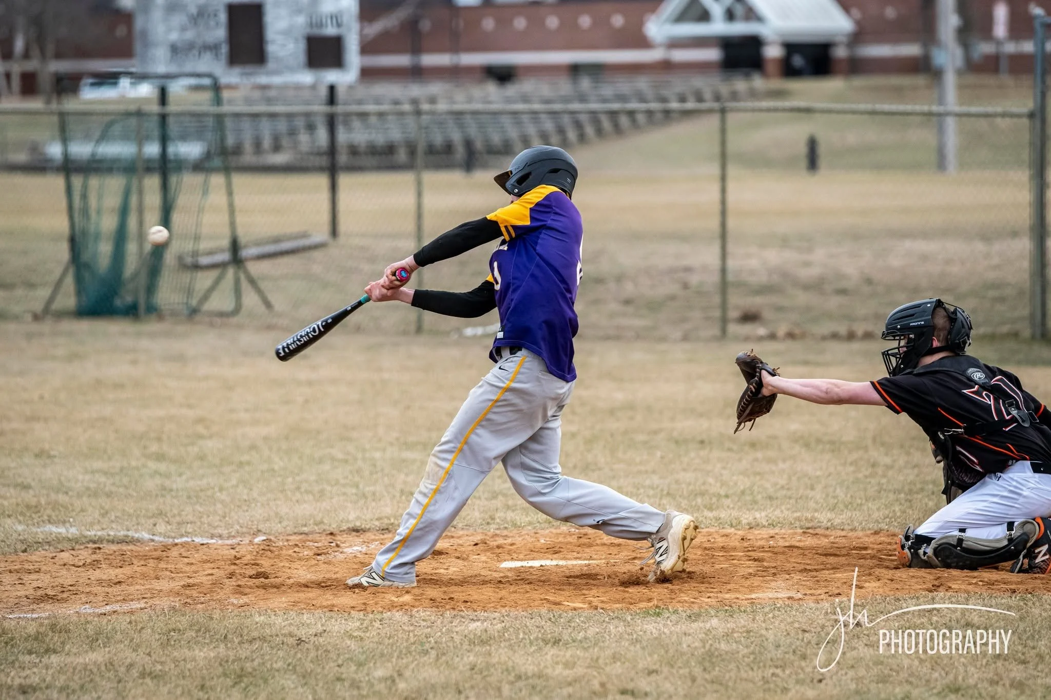 A young baseball player wearing a helmet and purple jersey is swinging a bat at a pitched ball on a baseball field.