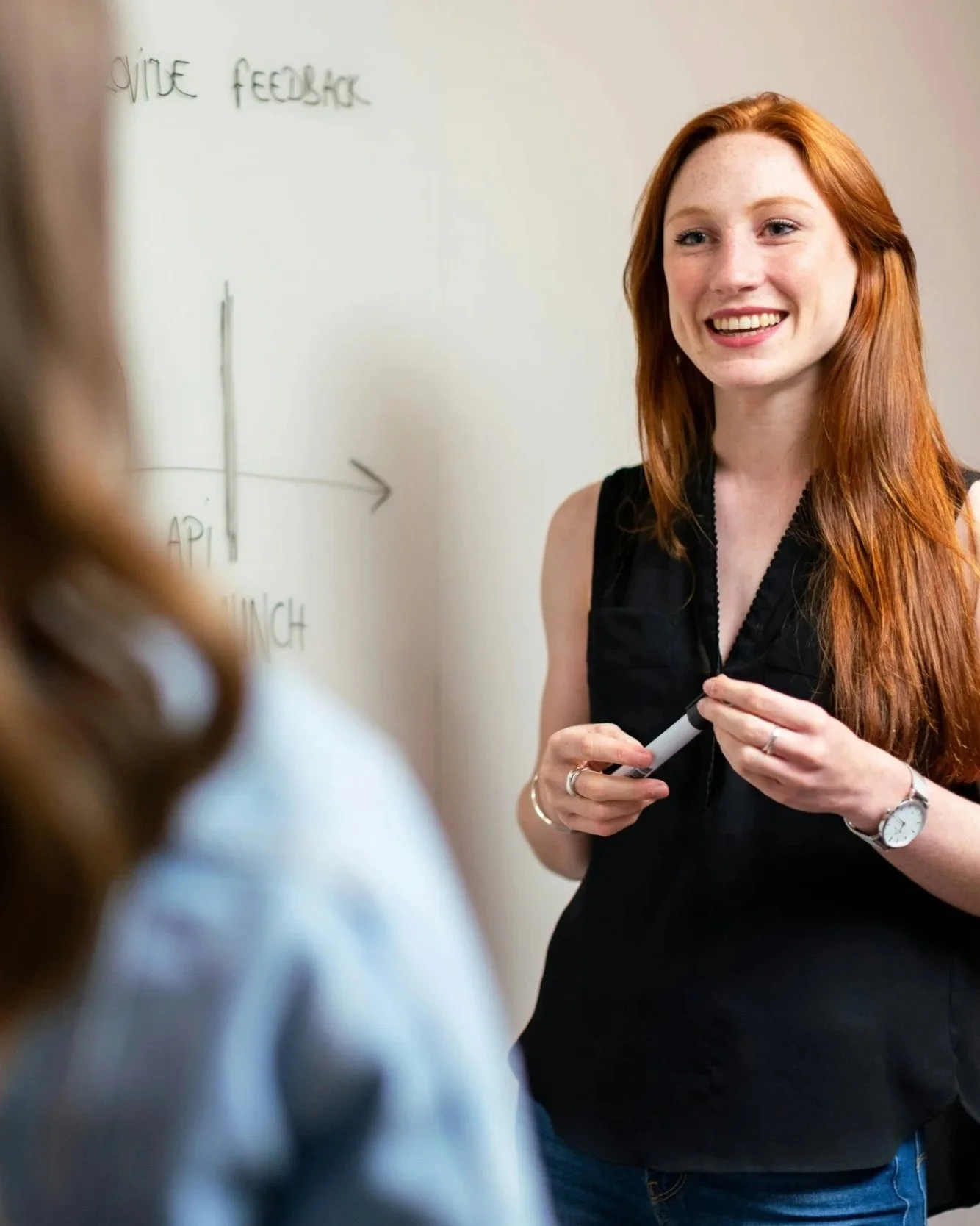 A woman with red hair, wearing a black sleeveless top, smiling and holding a white marker, standing in front of a whiteboard with handwritten notes.