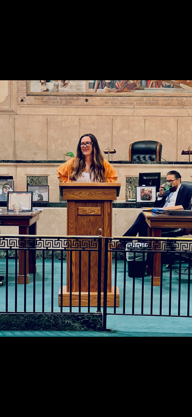 Woman speaking at podium in a formal setting, man sitting to her right drinking from a glass, framed photographs on tables, decorative metal railing in front, and murals on the wall behind.