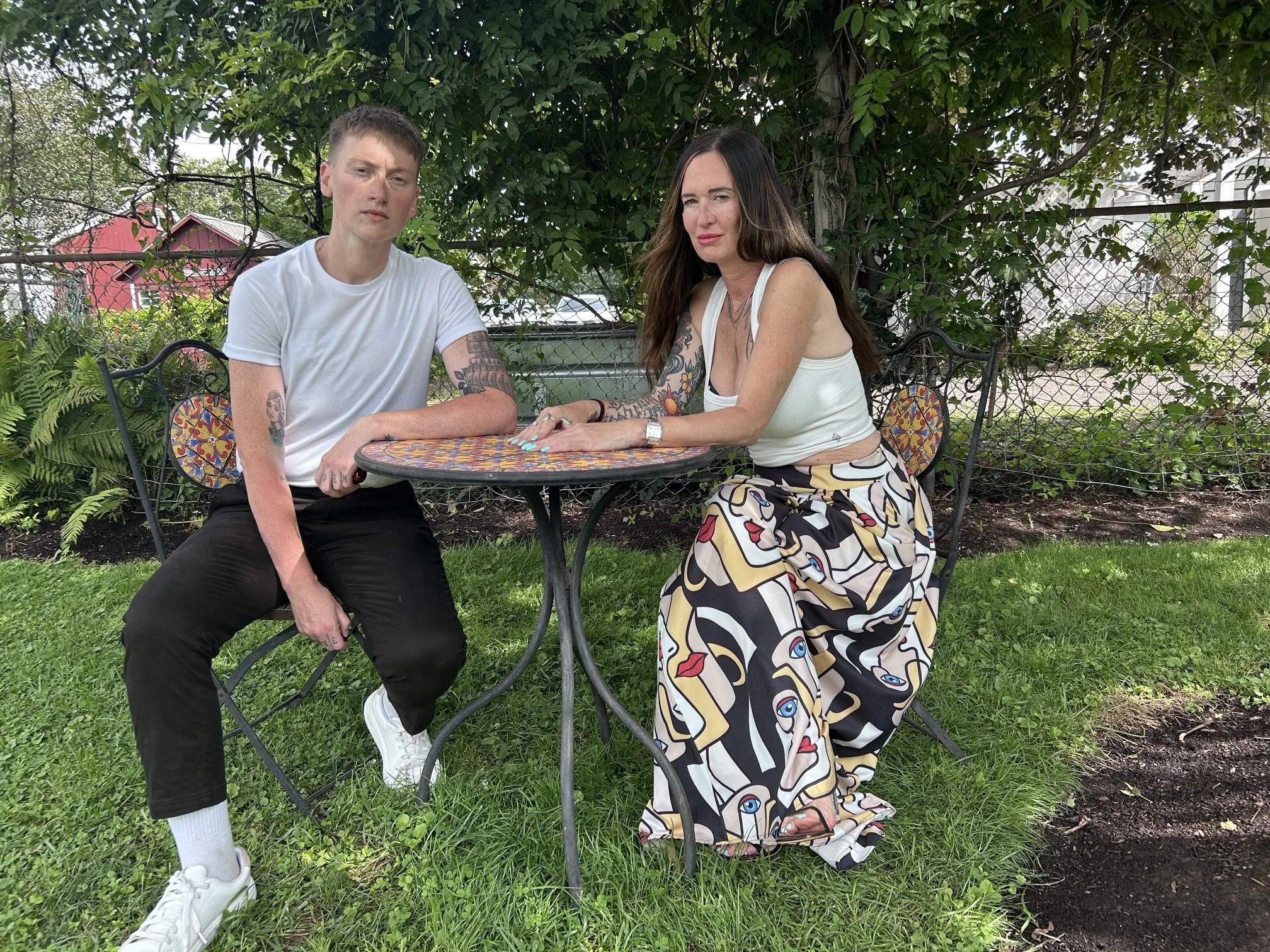Two women sitting at a colorful outdoor table with a lush green background, one wearing a white tank top and patterned pants, and the other in a white t-shirt and black pants, both with tattoos.