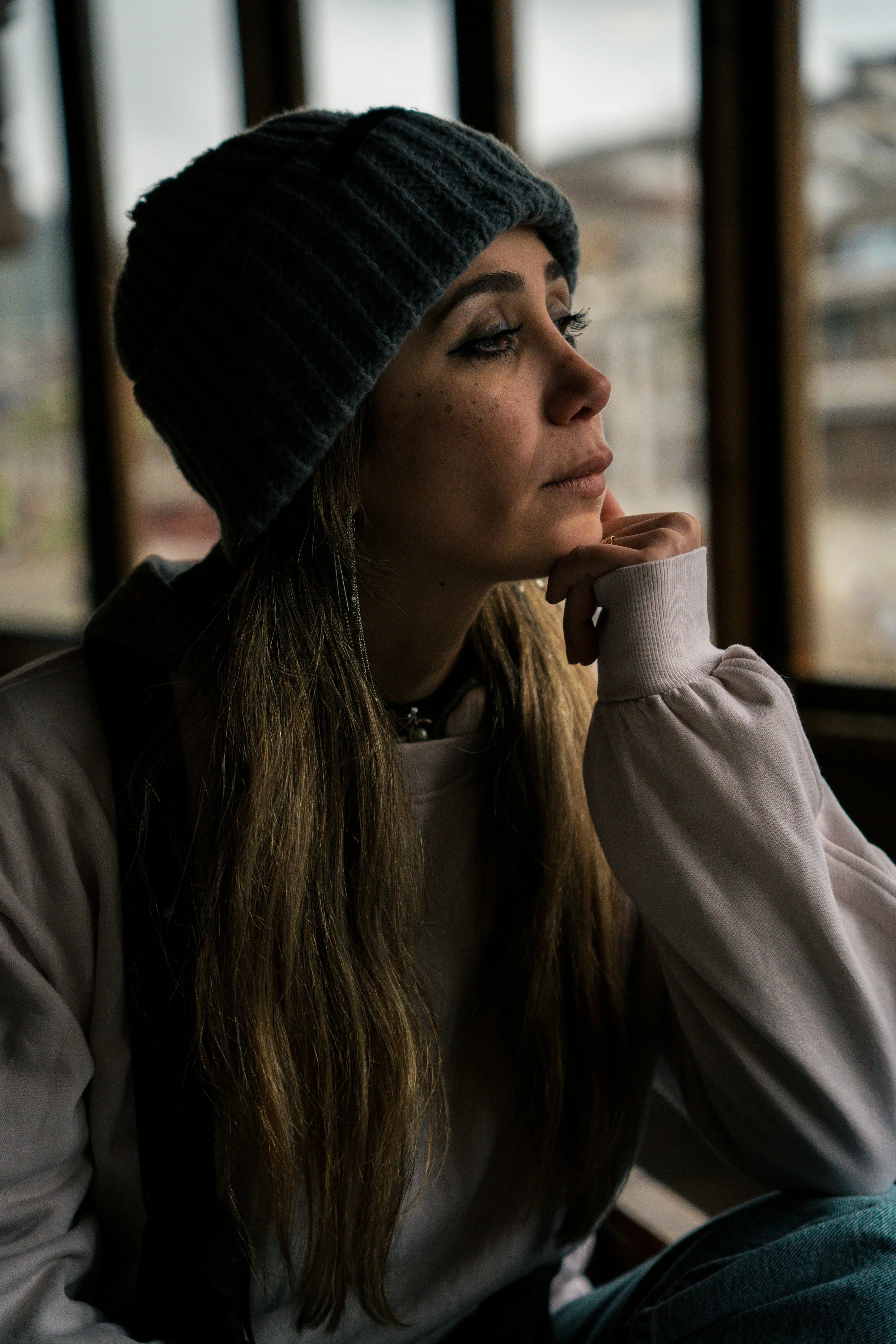 A young woman with long hair, wearing a dark knit beanie, sits indoors near a window, resting her chin on her hand and looking thoughtfully outside.