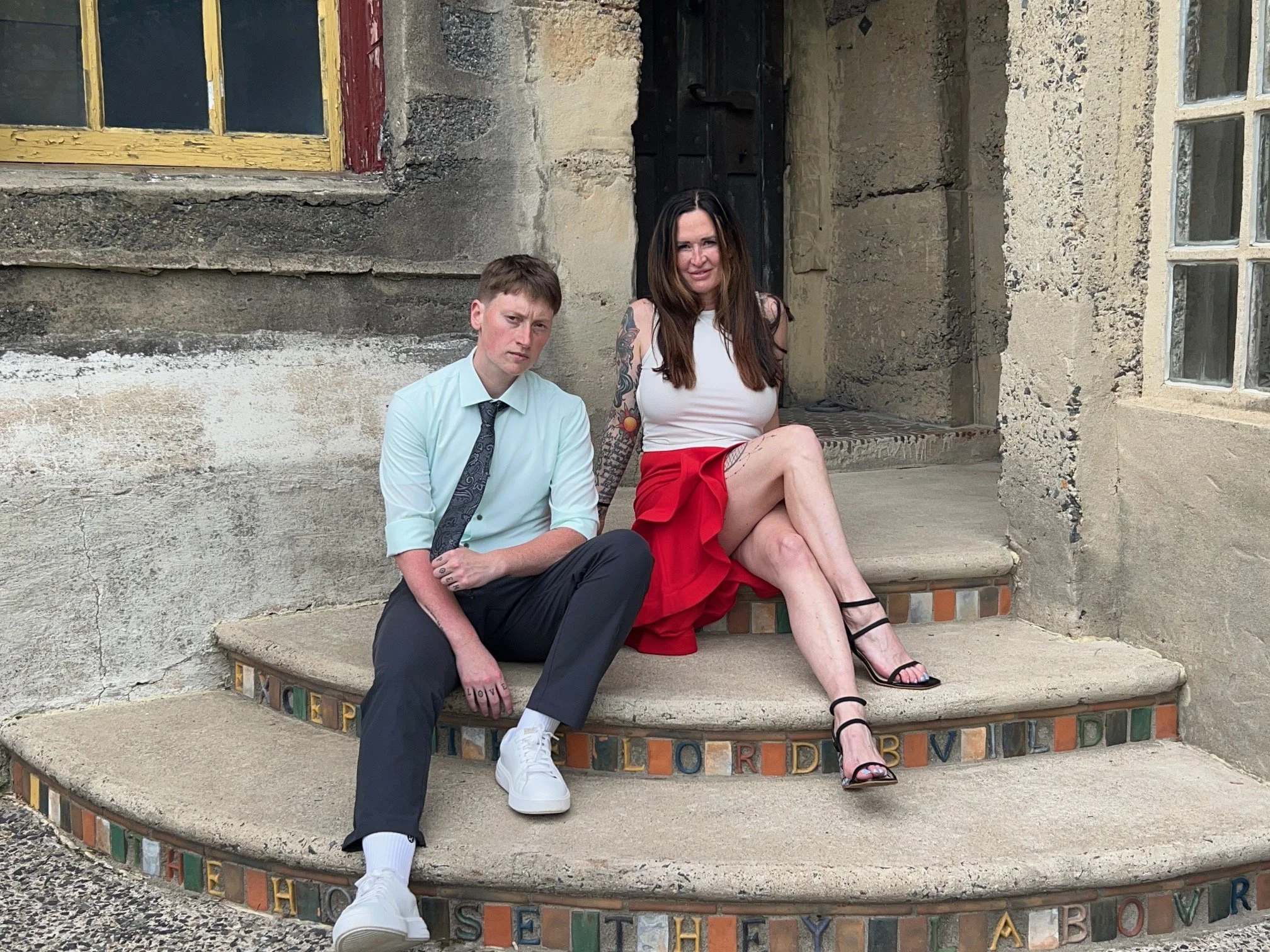 A man and a woman sitting on the steps of a rustic building with textured stone walls and colorful windows. The man is wearing a light blue shirt, black pants, and white sneakers, and has short brown hair. The woman is dressed in a white top and a red skirt, with long brown hair, black high-heeled sandals, and tattoos on her arms.