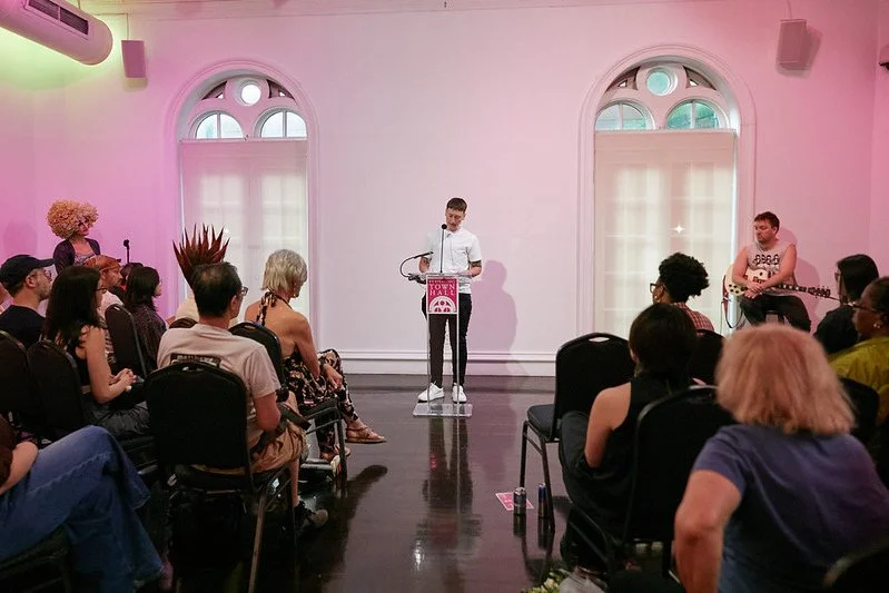 A young man is standing at a podium speaking to an audience in a bright room with pink lighting, arched windows, and a guitarist seated to his right.