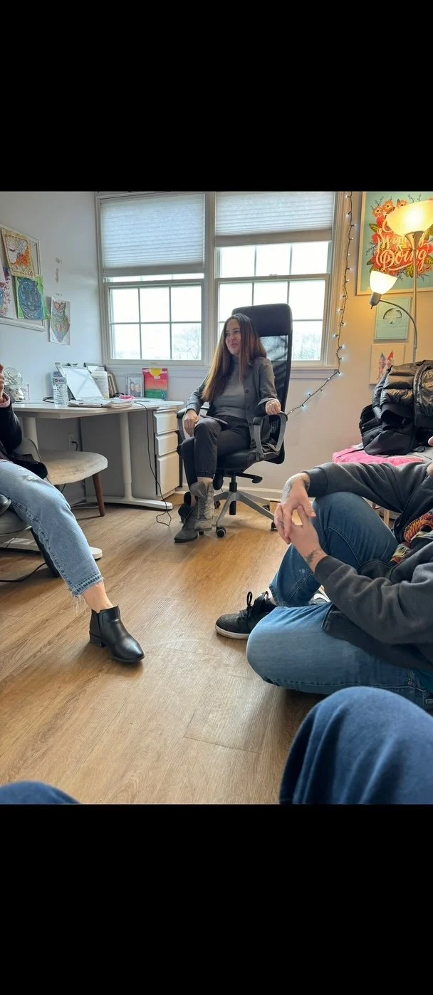 Three people sitting in a casual room with a woman sitting in a black office chair at the center, speaking to two others. The room has windows, artwork, and a lit floor lamp with string lights.