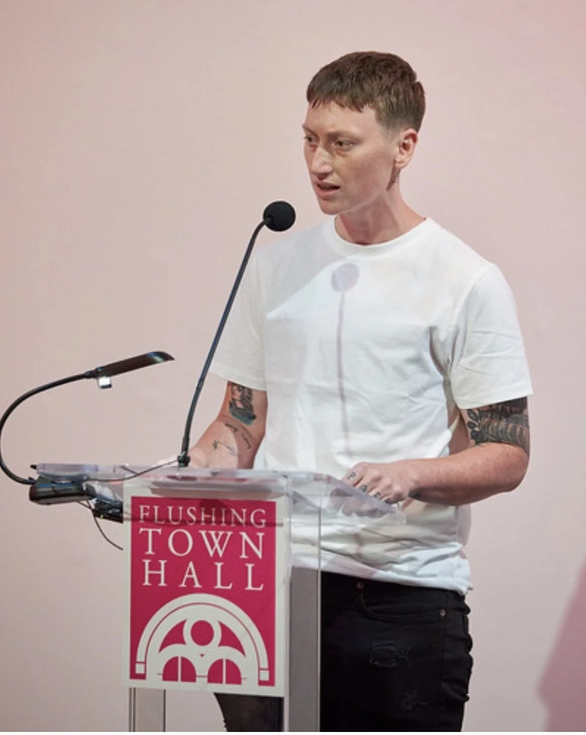 A person with short hair, tattoos on arms, wearing a white t-shirt, speaking at a town hall with a pink sign that reads "Flushing Town Hall."