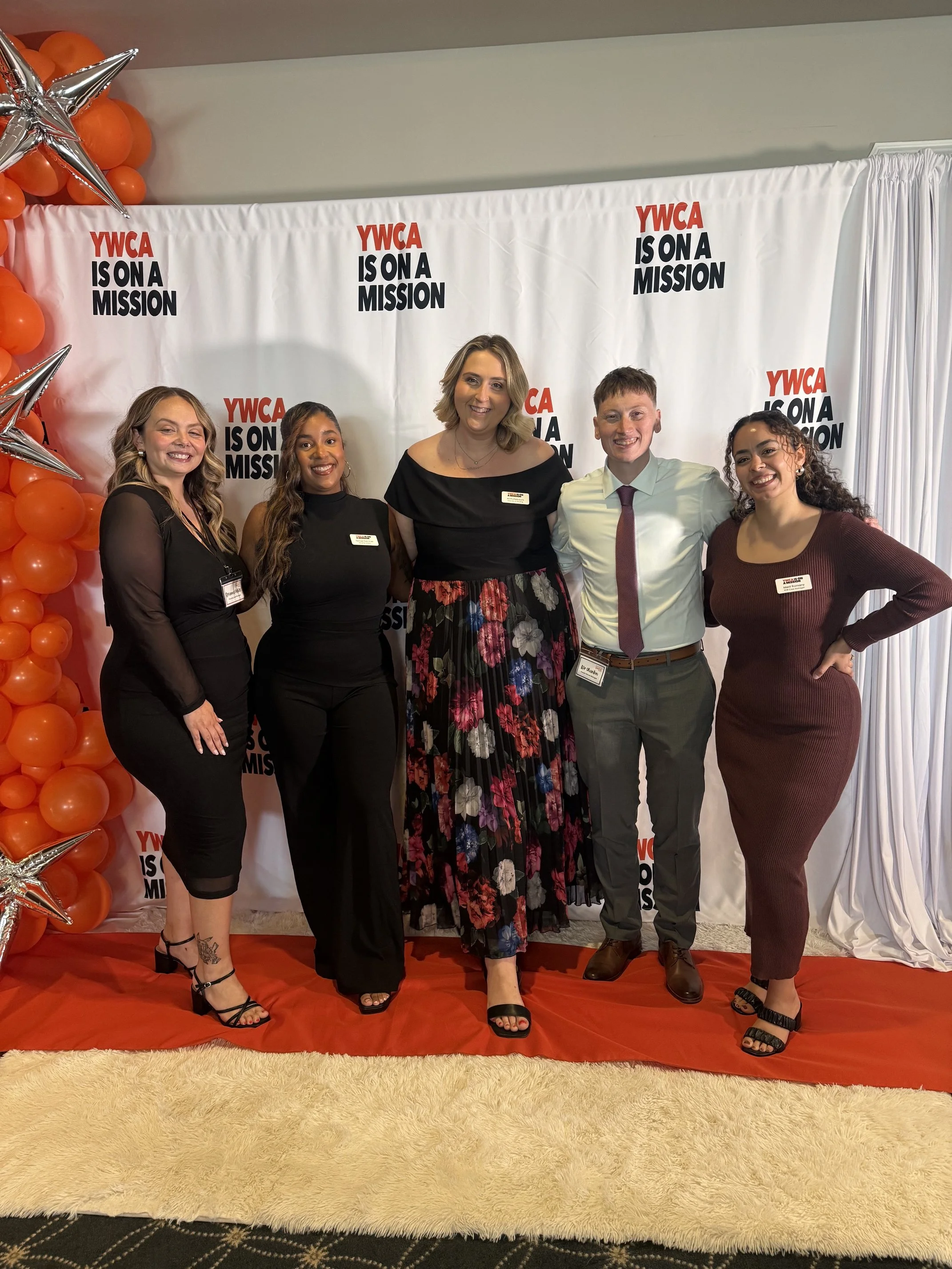 Group of five people standing in front of a YWCA backdrop decorated with orange balloons and silver stars, wearing formal attire at an event.