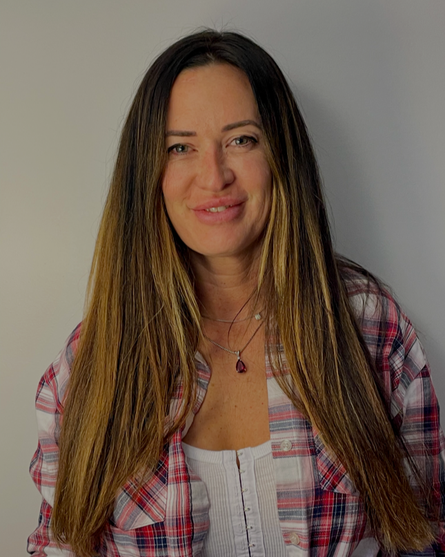 A woman with long brown hair wearing a plaid shirt and layered necklaces, smiling softly against a plain light-colored wall.