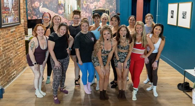 A group of fifteen women posing together in an art gallery with colorful paintings and blue walls.