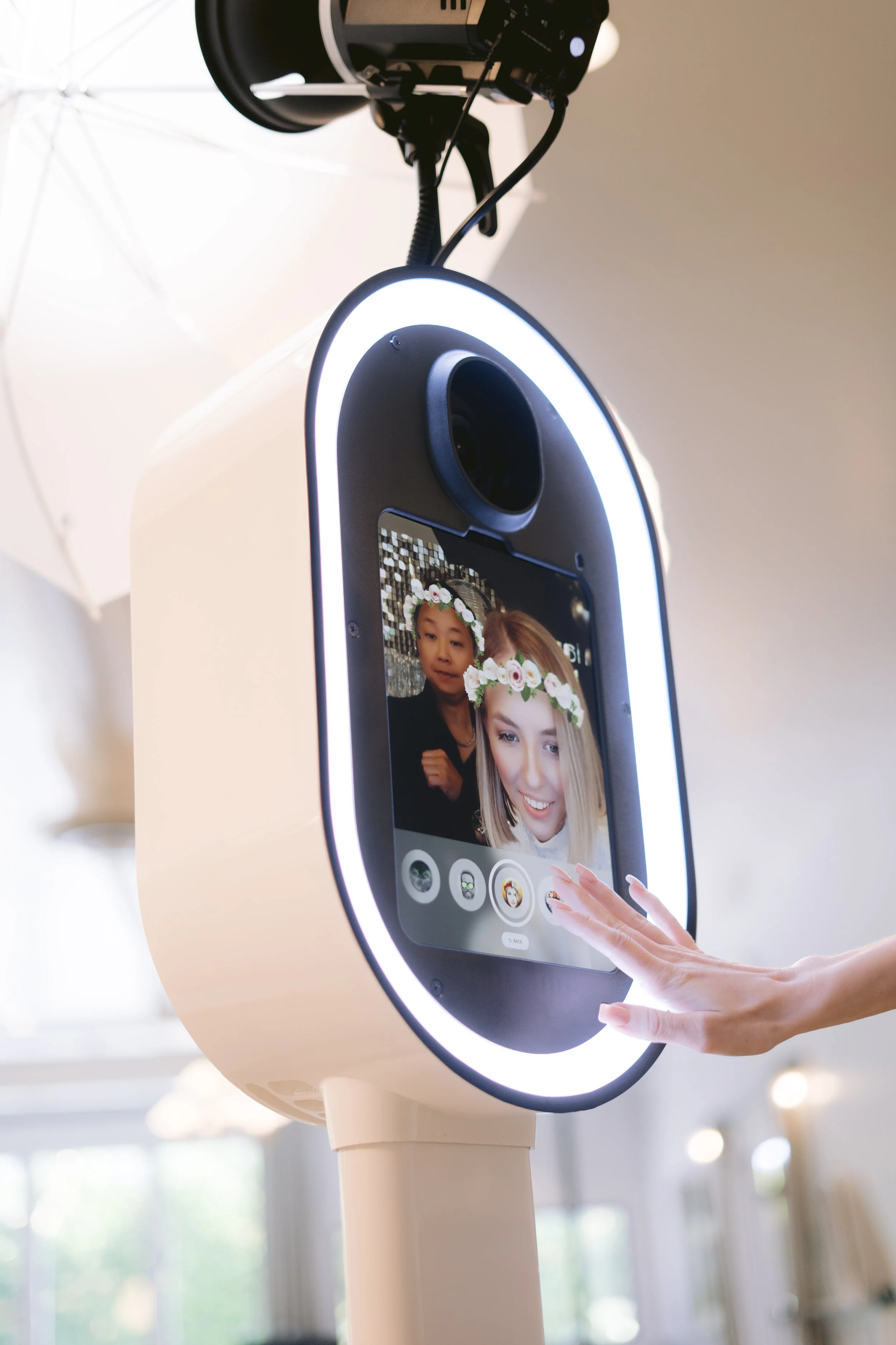 A person touching a digital kiosk screen with a photo of two women, one with blonde hair and wearing a flower crown, and another with dark hair, in an indoor setting with a white umbrella light in the background.