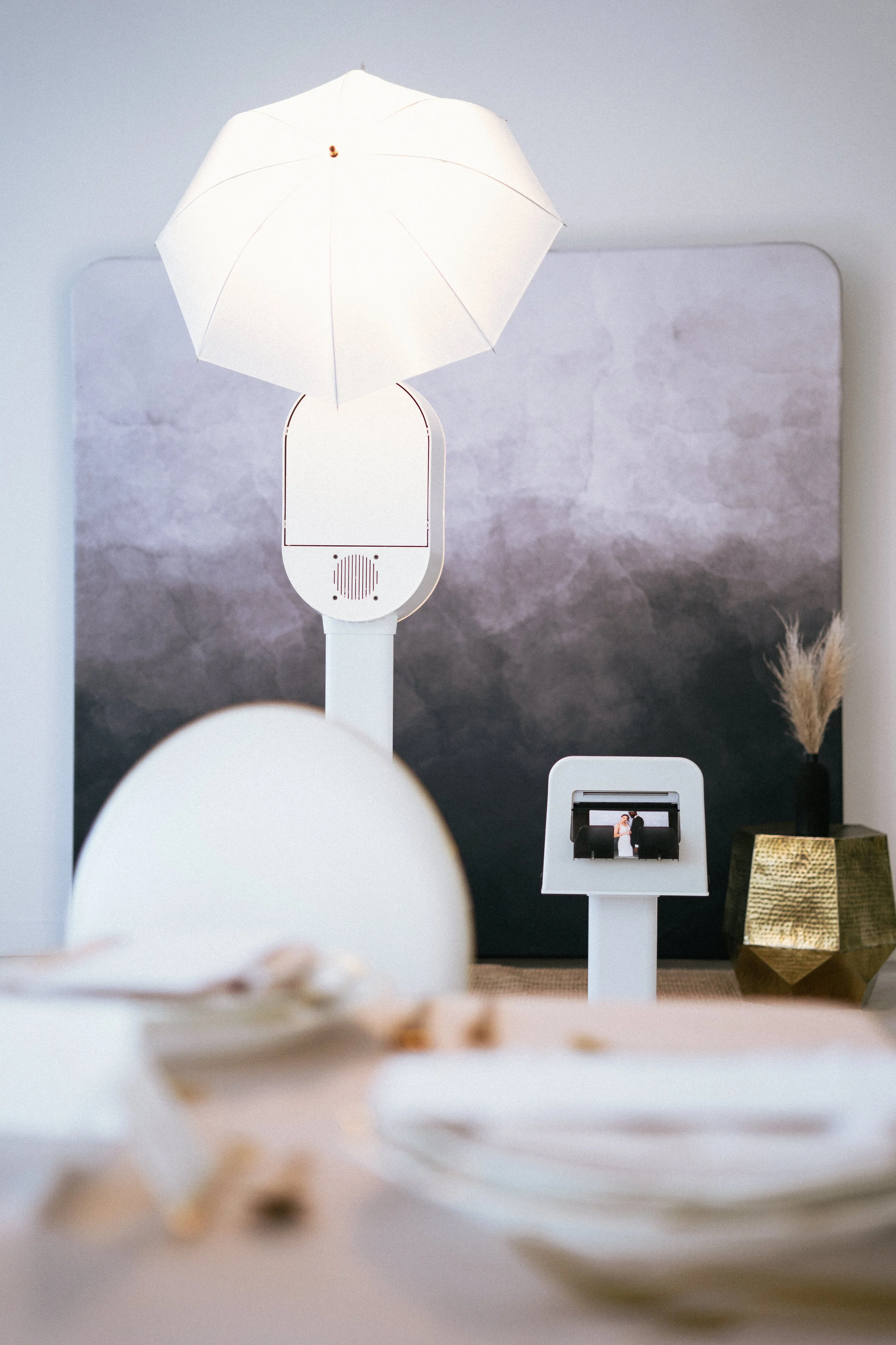 Photo of a modern digital photo booth with a screen showing a couple taking a selfie, positioned in a stylish room with a textured gray wall, a large white umbrella-style lamp, and a decorative gold object in the background.