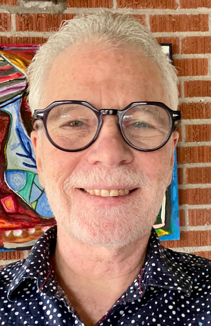 Headshot of Gregory Stock standing in front of a brick wall with colorful artwork.