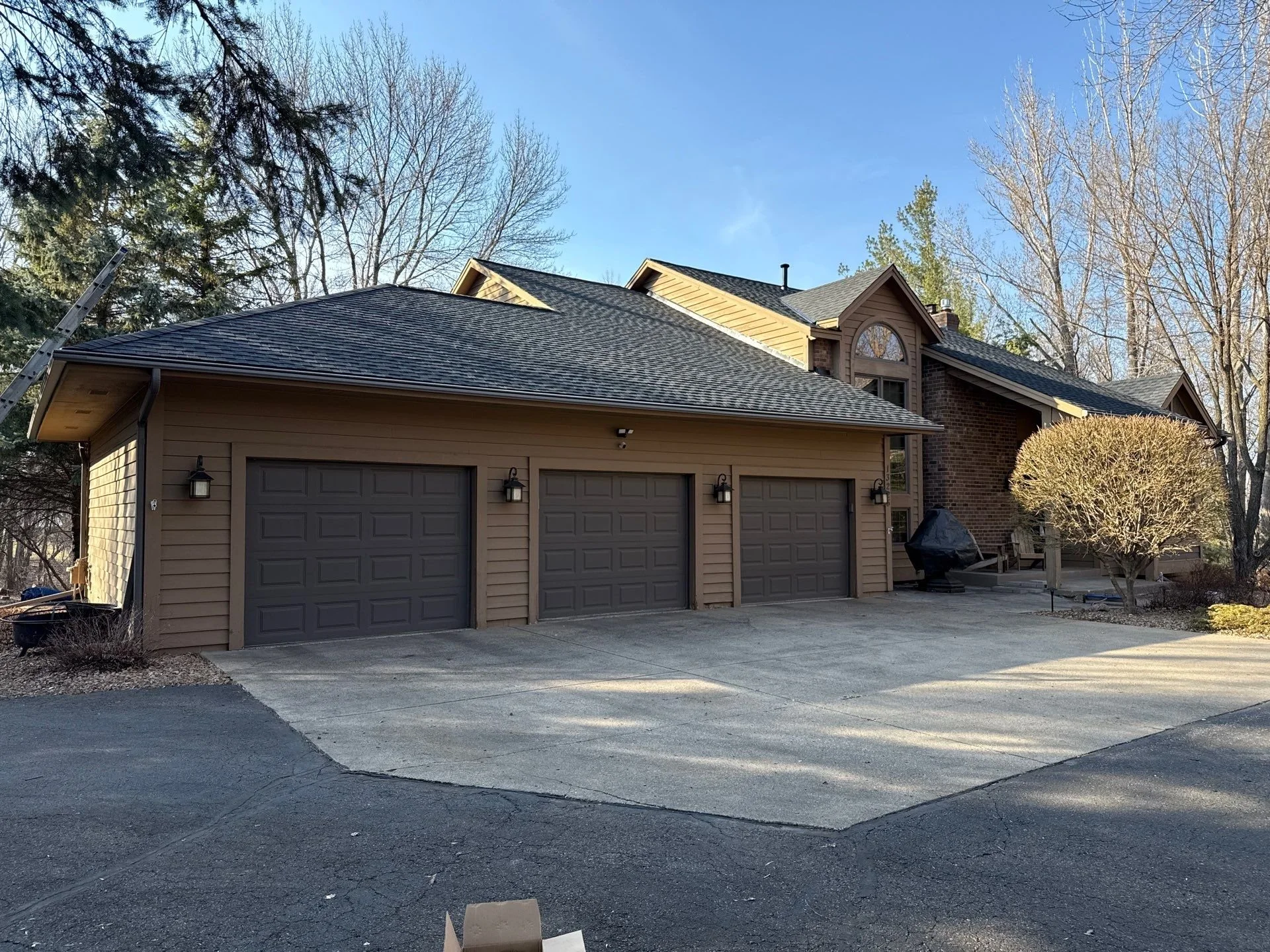 A residential house with a three-car garage, beige siding, and a wooden deck. The house has a gable roof and brick chimney, surrounded by leafless trees under a clear blue sky.