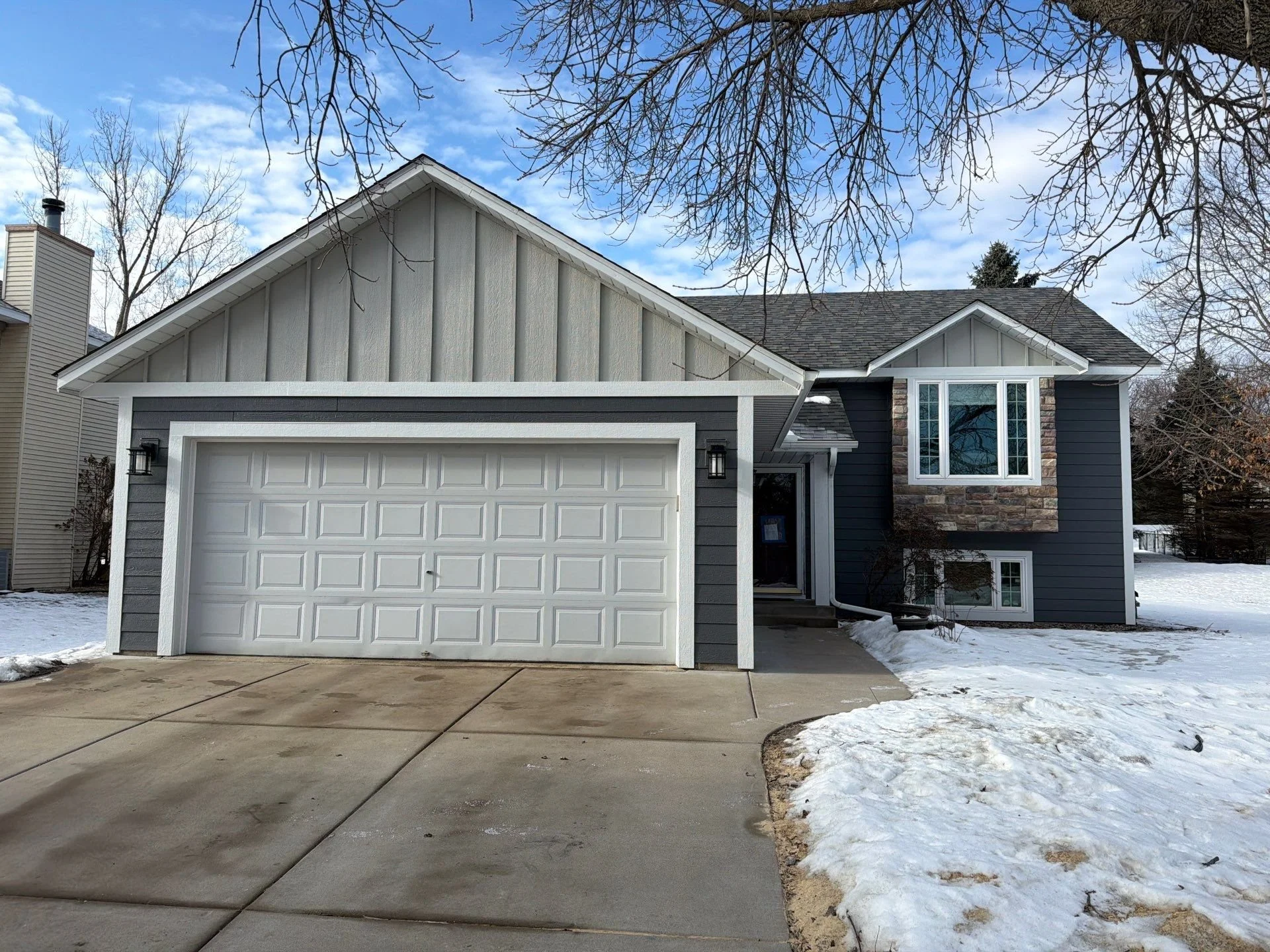 Front view of a modern two-story house with a garage, dark gray siding, and stone accents, with snow on the ground and leafless trees in the background.