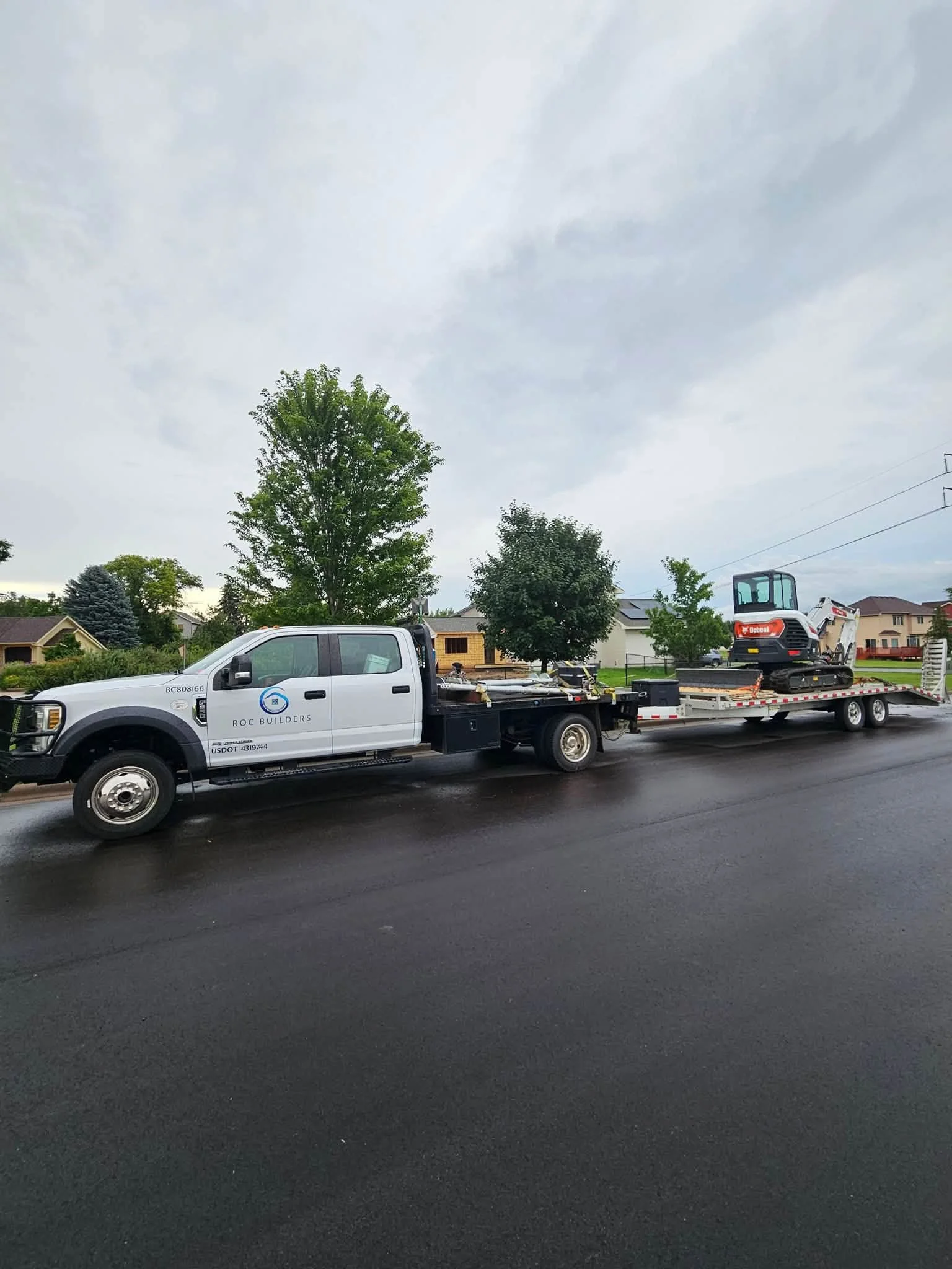 A white flatbed truck with a logo and company name parked on the street. The truck is carrying construction equipment, including an excavator, with a residential neighborhood and trees in the background under a cloudy sky.