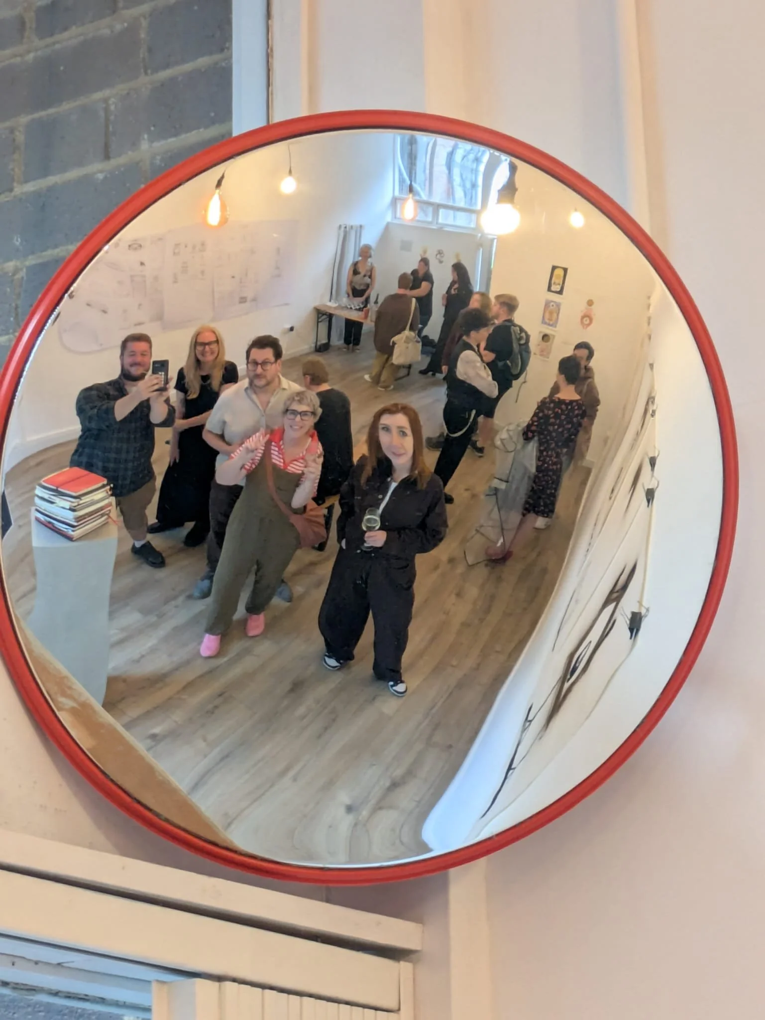 A group of people gathering and taking photos in a room with artwork and posters on the walls, reflected in a large circular mirror with a red border.