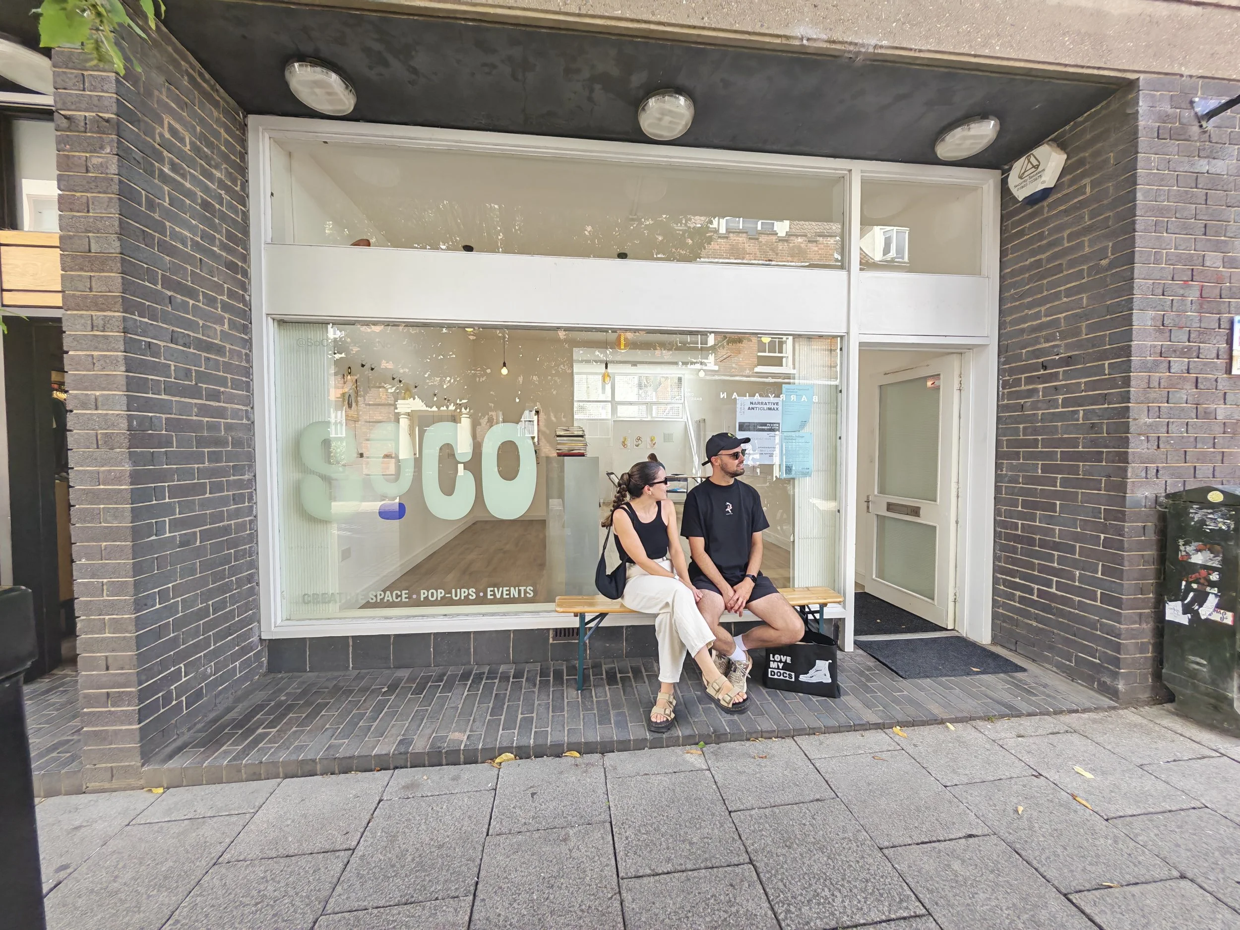 A storefront with a large glass window and a white door, two people sitting on a bench outside, and a sign promoting space, pop-ups, and events inside. The building has dark brick walls on either side of the entrance, with three flush-mounted lights 