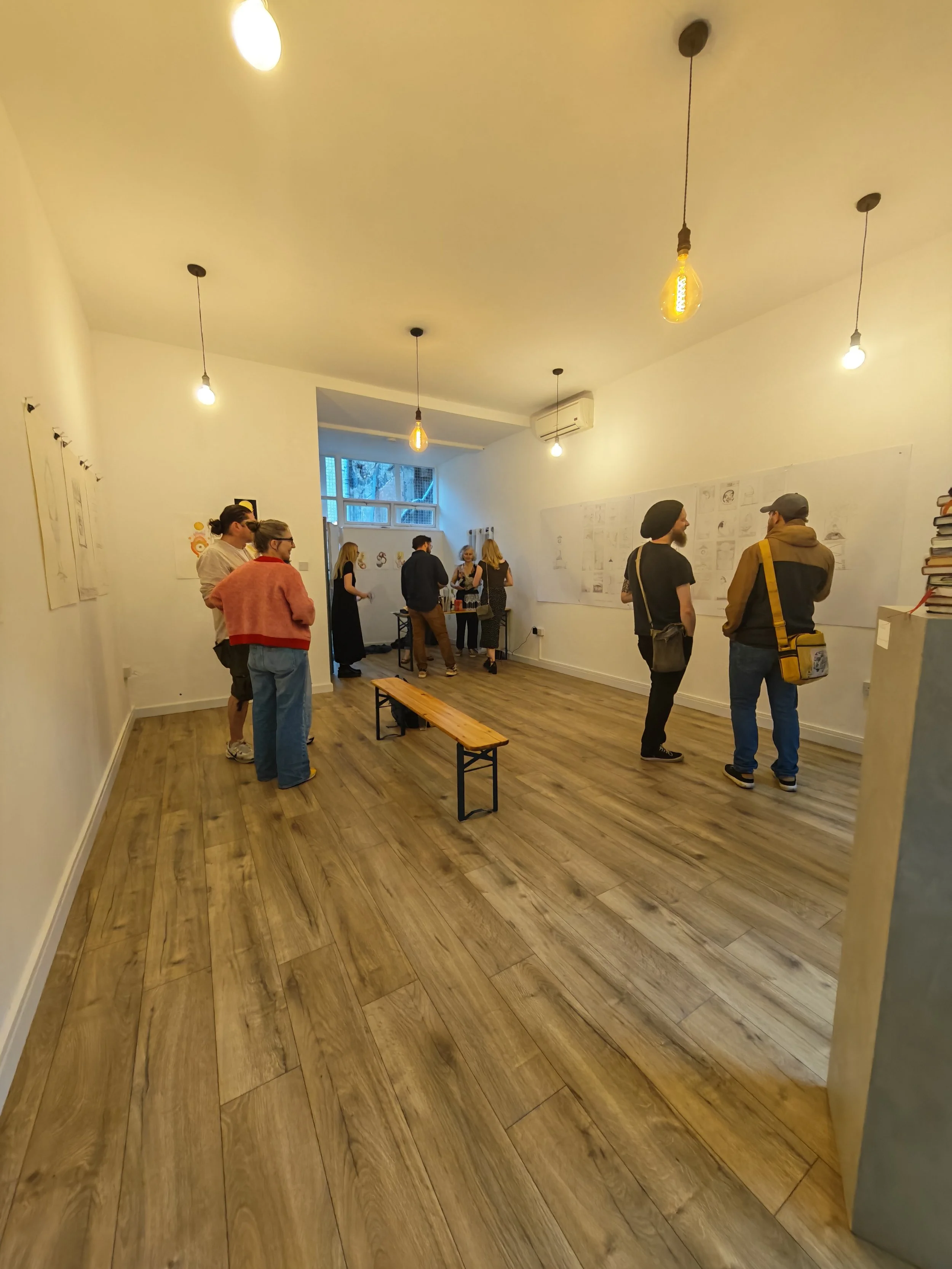 People gather in an art gallery or exhibition space, viewing drawings and sketches displayed on white walls, with warm lighting and wood flooring.