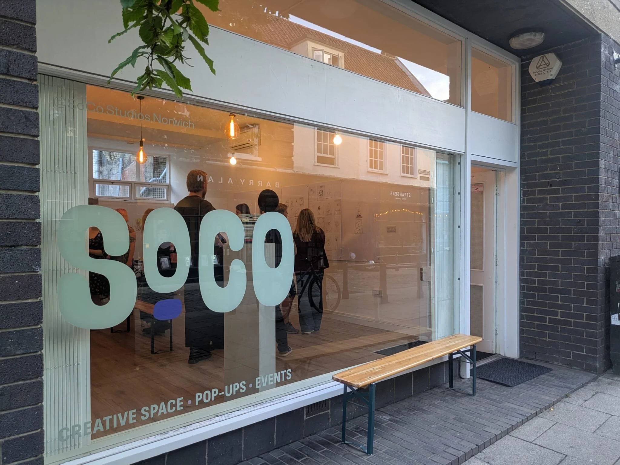 The exterior of a storefront with large glass windows showing a cozy interior with hanging lights and people inside. The window has text promoting creative space, pop-ups, events, and more. A small wooden bench is placed outside on the sidewalk.