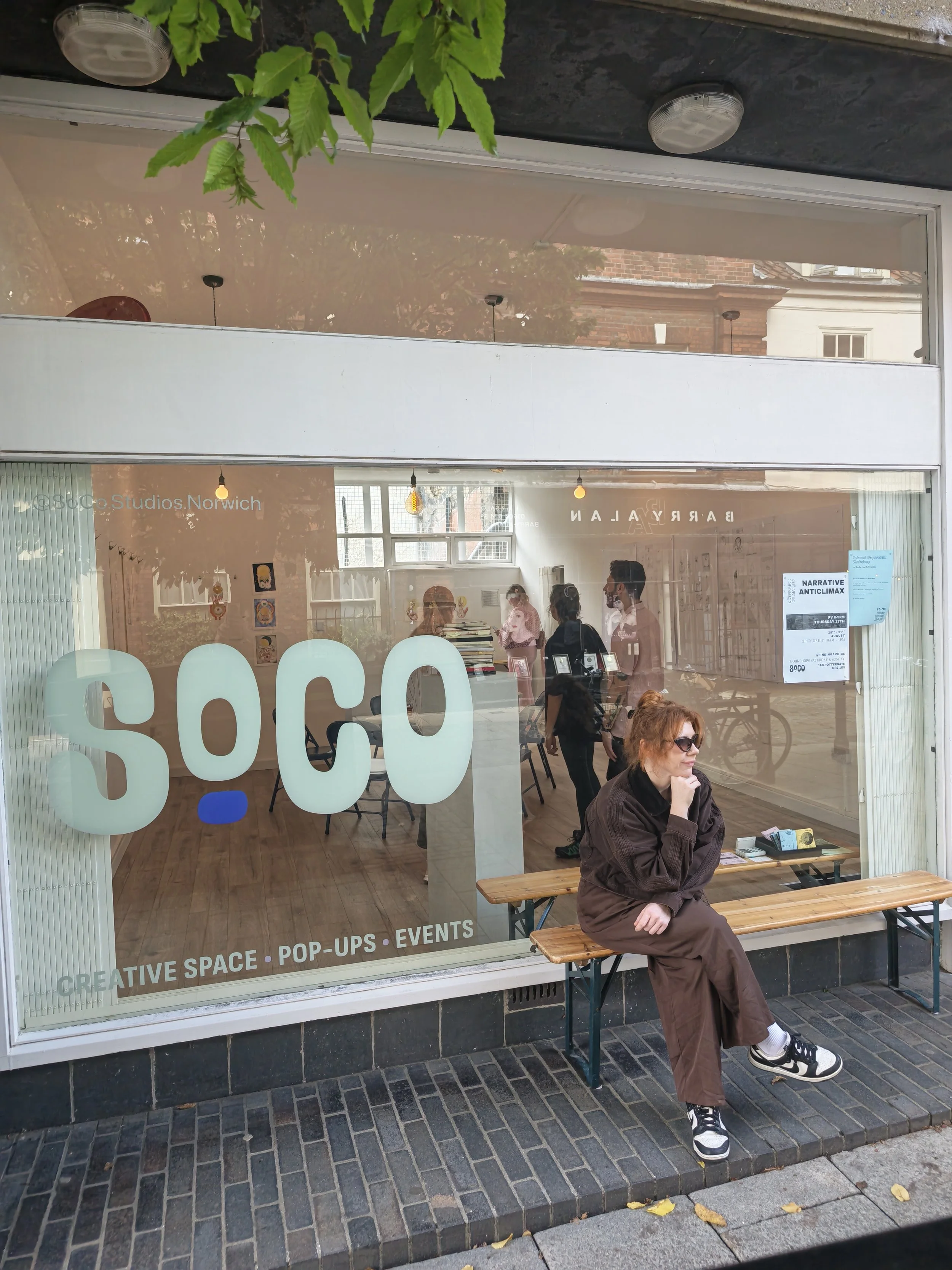 A woman with red hair and sunglasses sitting on a wooden bench outside a storefront, looking to the right. The storefront has large glass windows and door with white lettering for 'SOCIAL' and words 'CREATIVE SPACE,' 'POP-UPS,' and 'EVENTS' visible o
